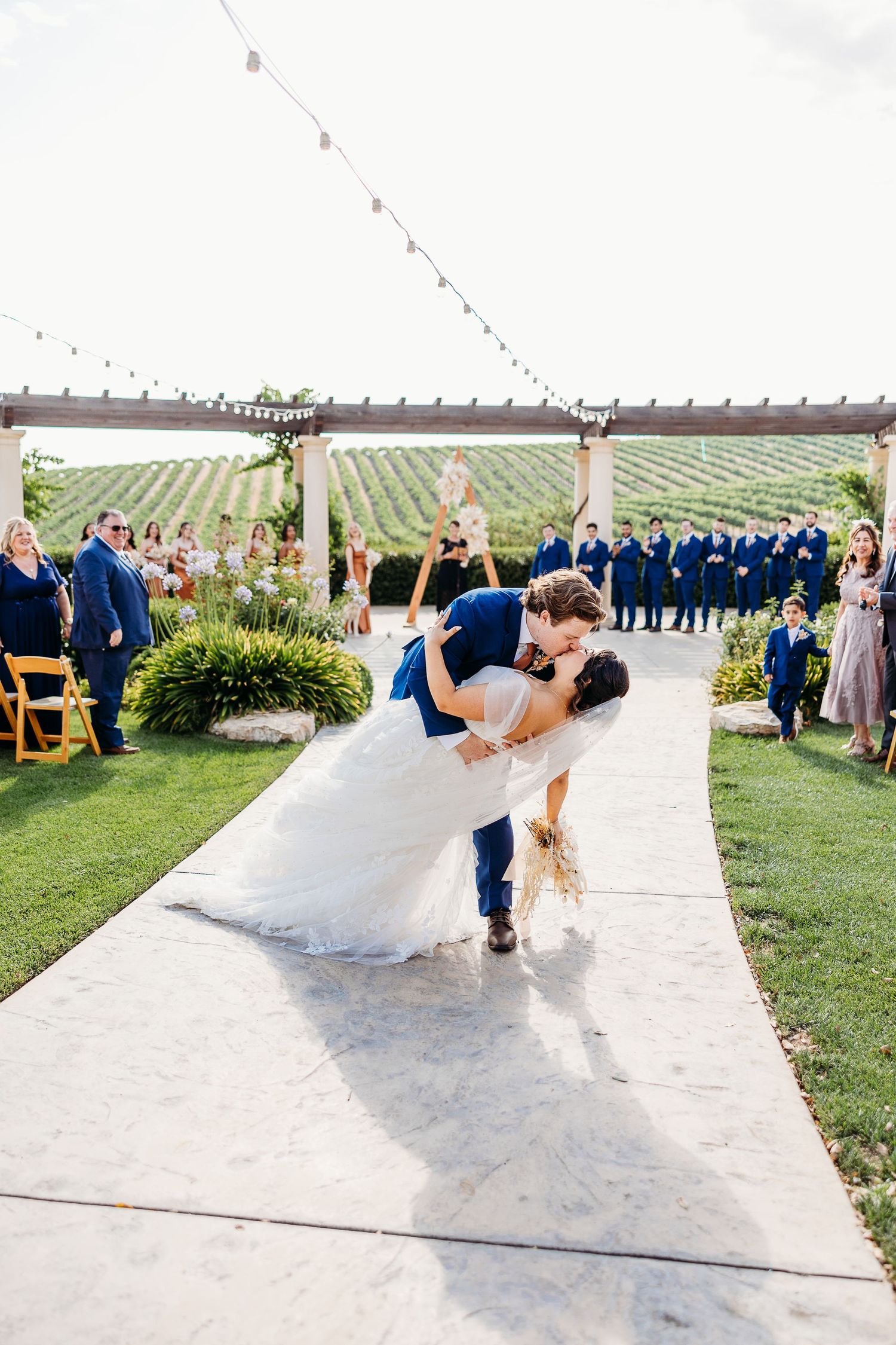 Romantic dip kiss captured during wedding celebration with guests cheering in background.