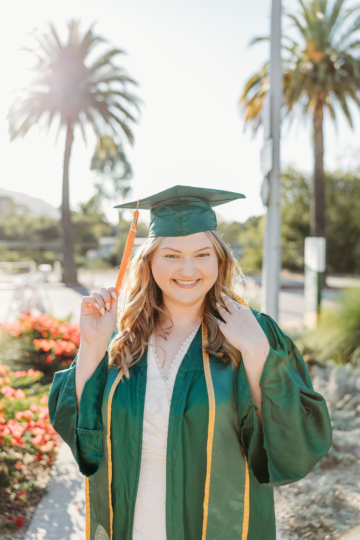 A graduate in emerald green cap and gown poses with palm trees and colorful flowers in the background.