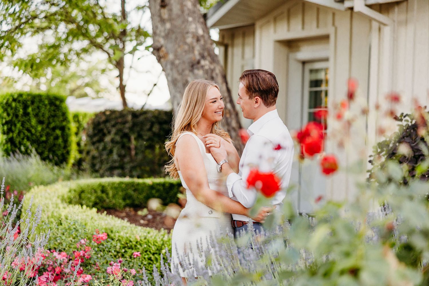 A couple shares a joyful moment together in a garden with blooming red flowers and a rustic house backdrop.