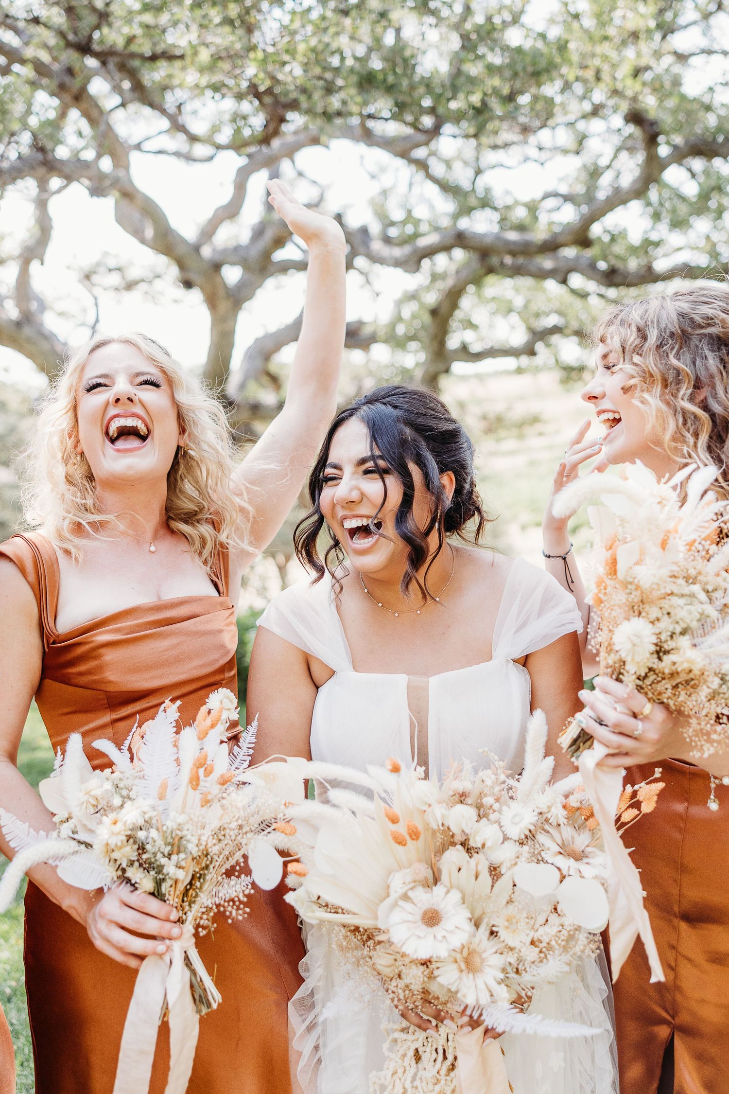 A joyful candid moment between bride and bridesmaids holding dried flower bouquets under oak trees.