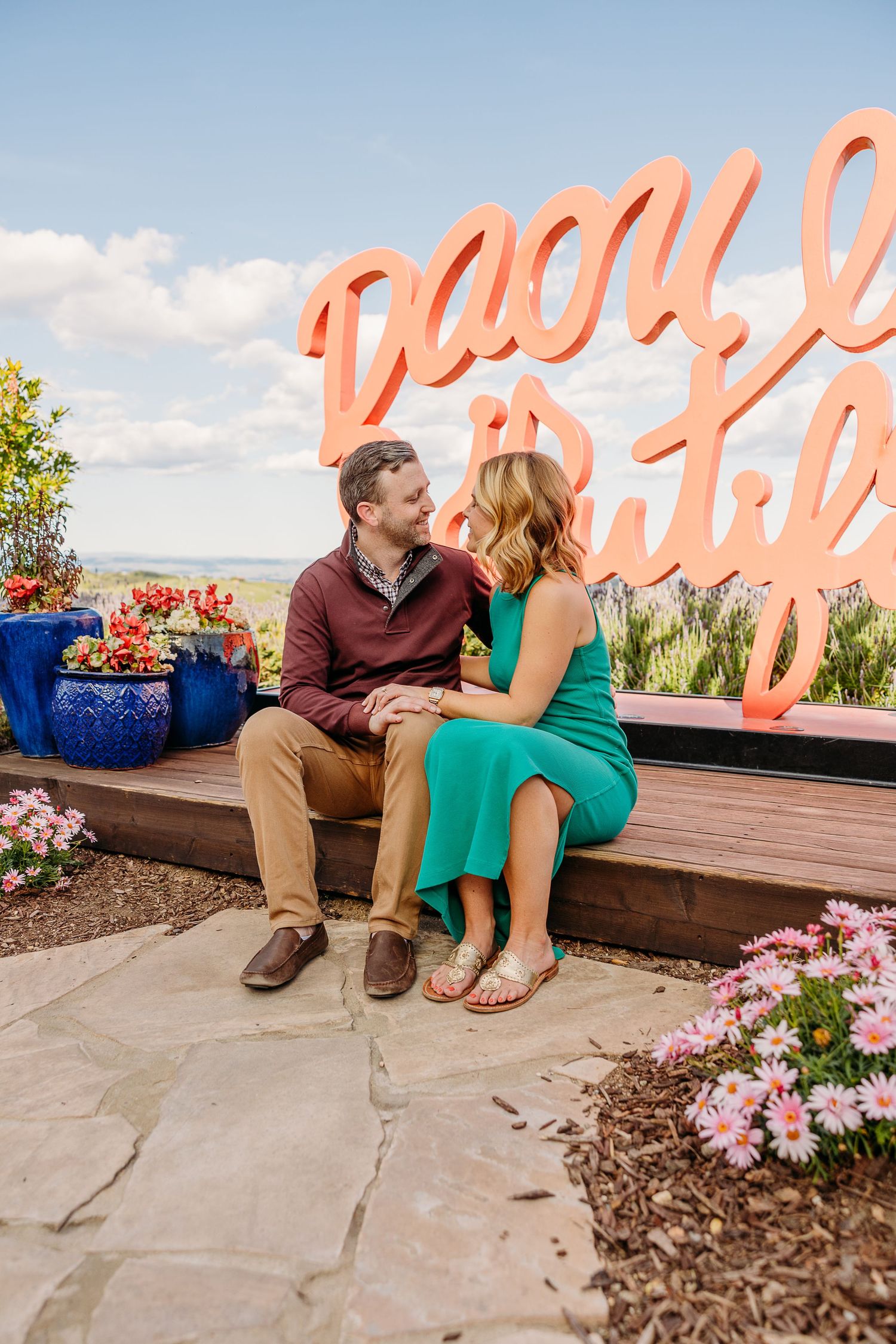 A couple sits together on a wooden bench against a scenic mountain backdrop with pink neon signage.