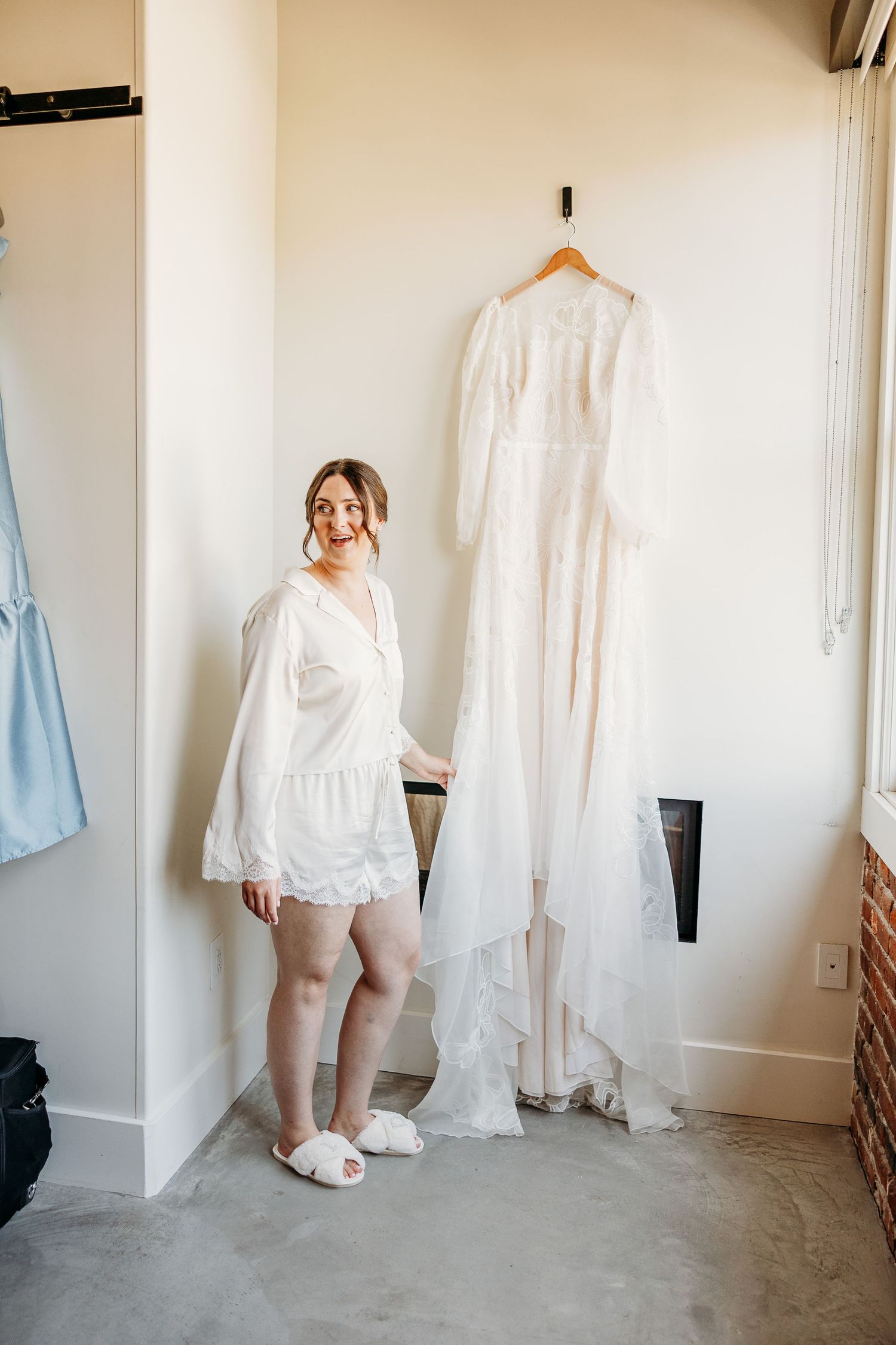 A white wedding dress hangs on display while someone in a white robe stands nearby.