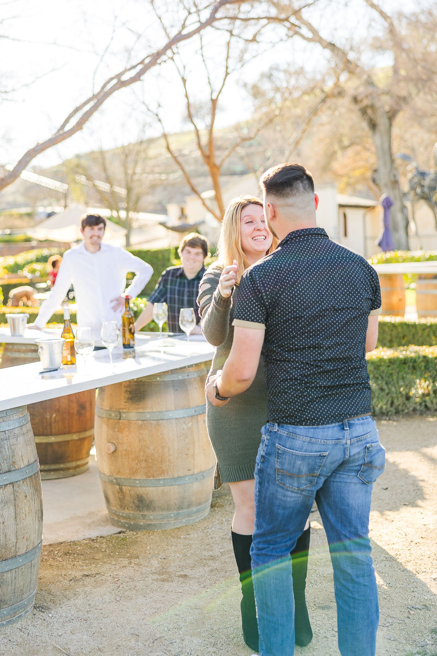 A romantic scene unfolds between two people at a rustic wine tasting setup with oak barrels.