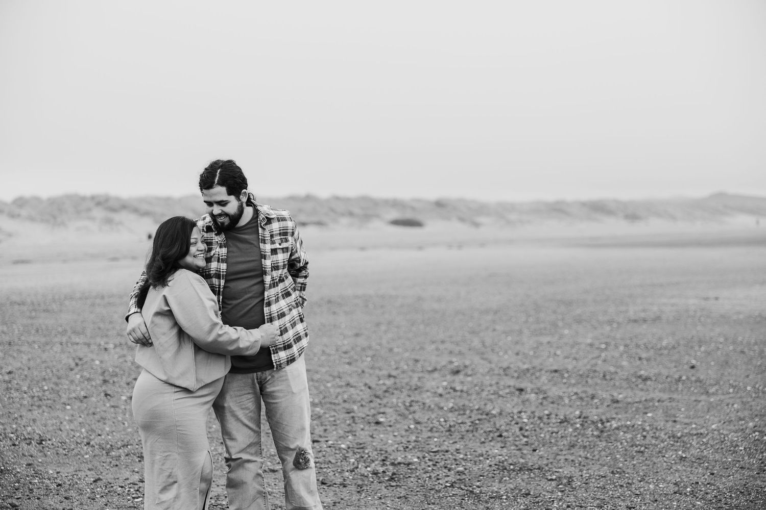 Black and white photo of an intimate couple embracing in an open field with rolling hills in the background.