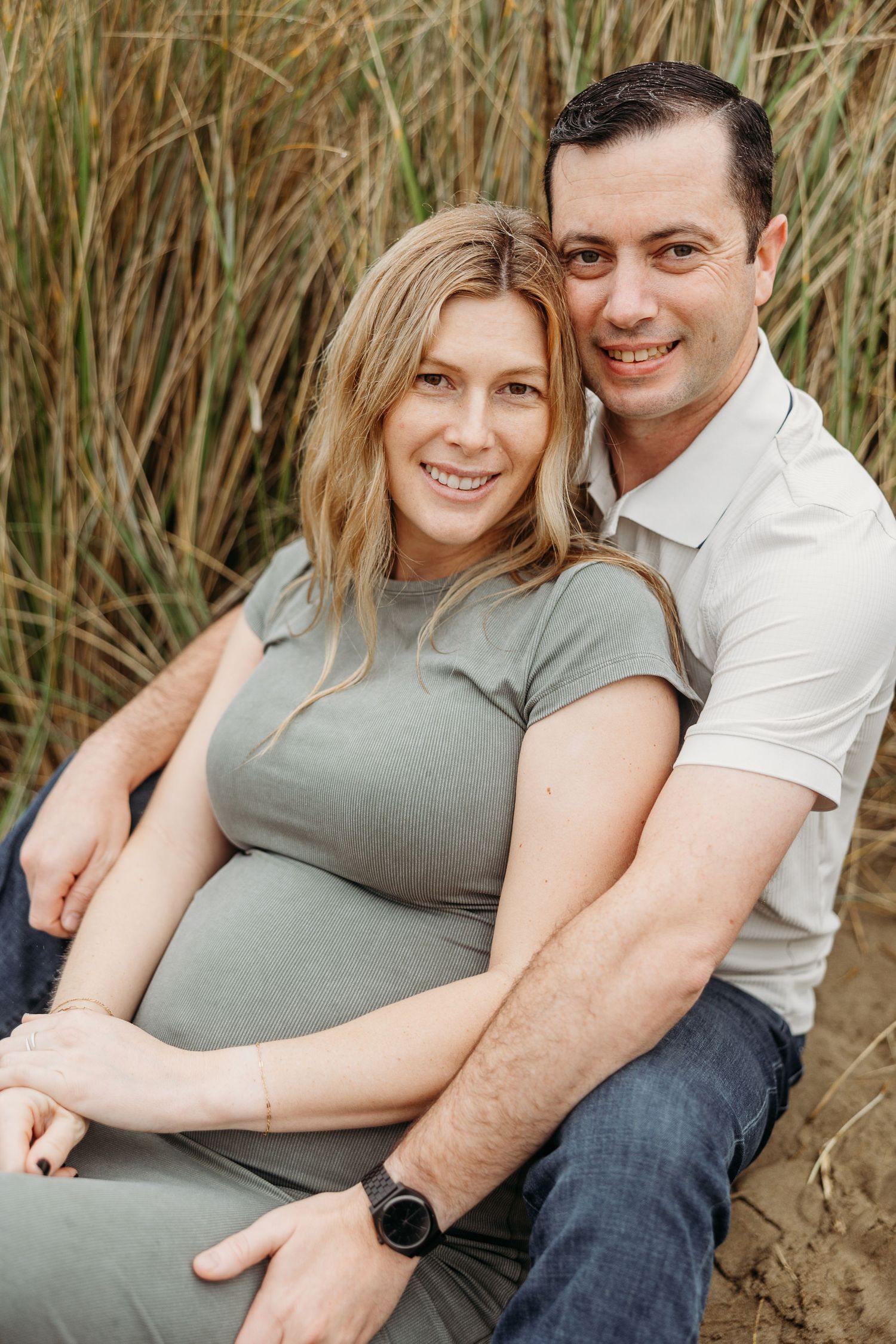 Intimate portrait of couple sitting together against beach grass backdrop during pregnancy photoshoot.