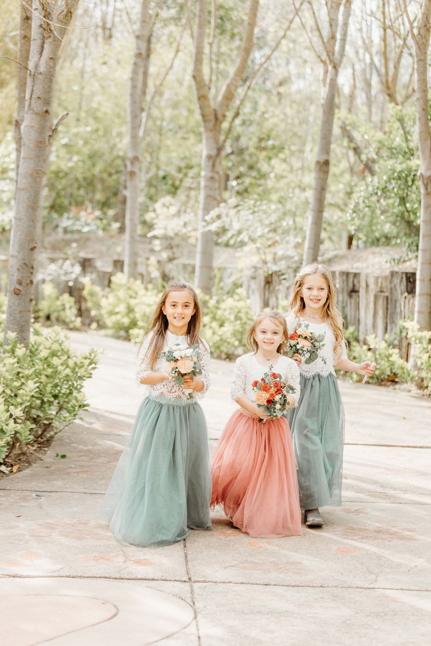 Three young flower girls in pastel tulle dresses walk down a garden path holding flower bouquets.