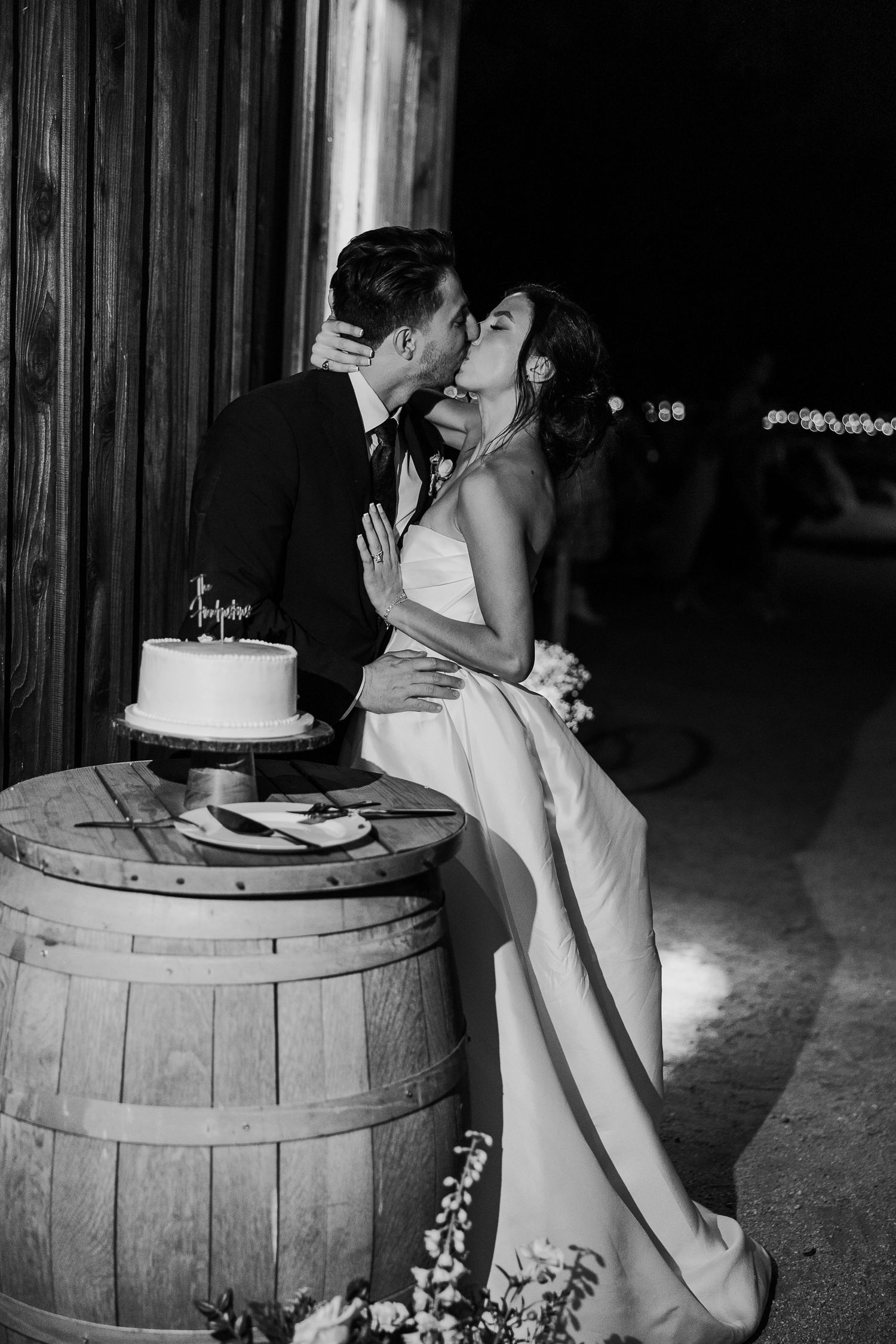 Romantic black and white photo of couple sharing intimate moment by wedding cake on wine barrel.