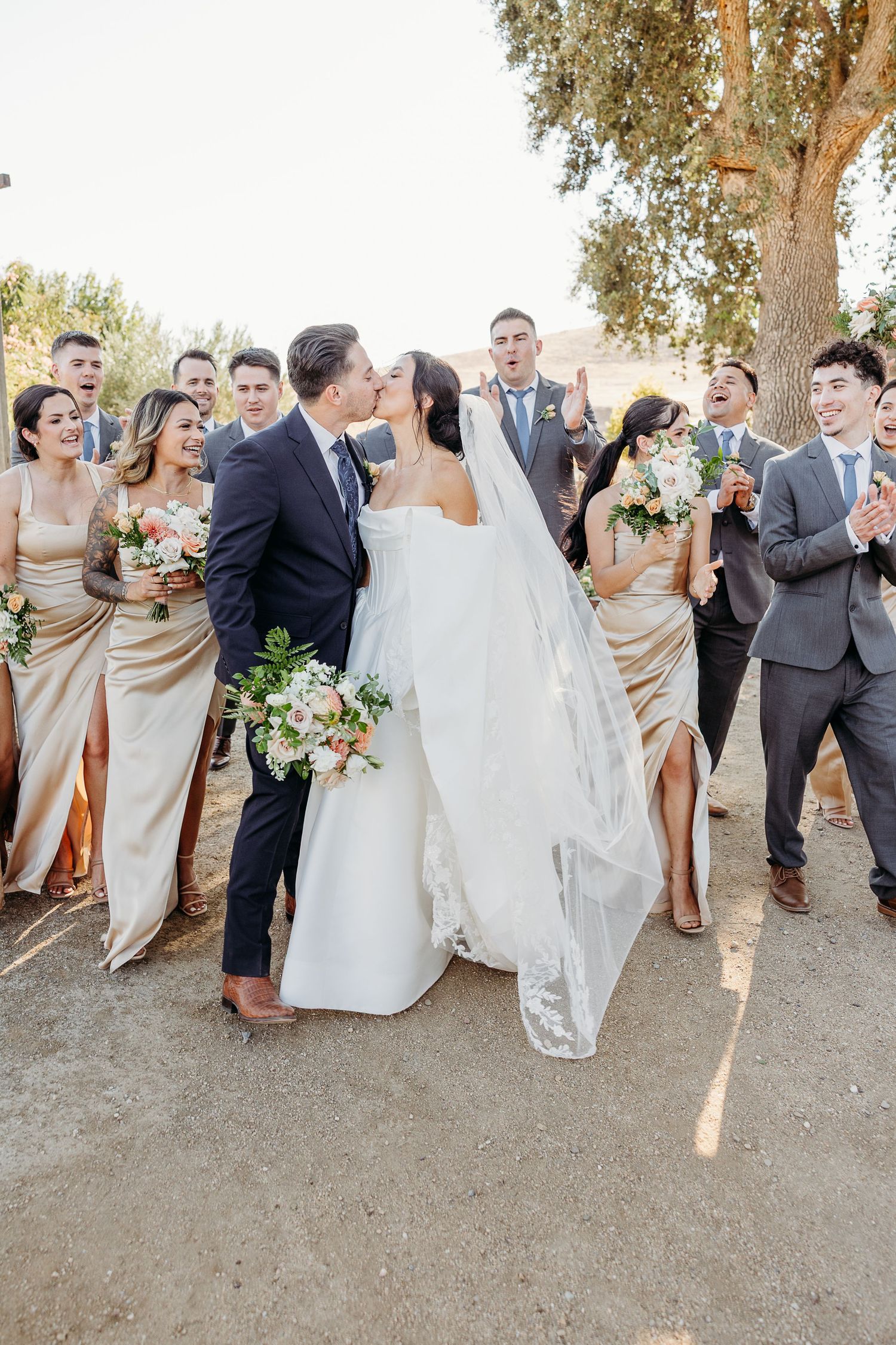 Wedding party in champagne bridesmaid dresses and gray suits pose together outdoors during golden hour.