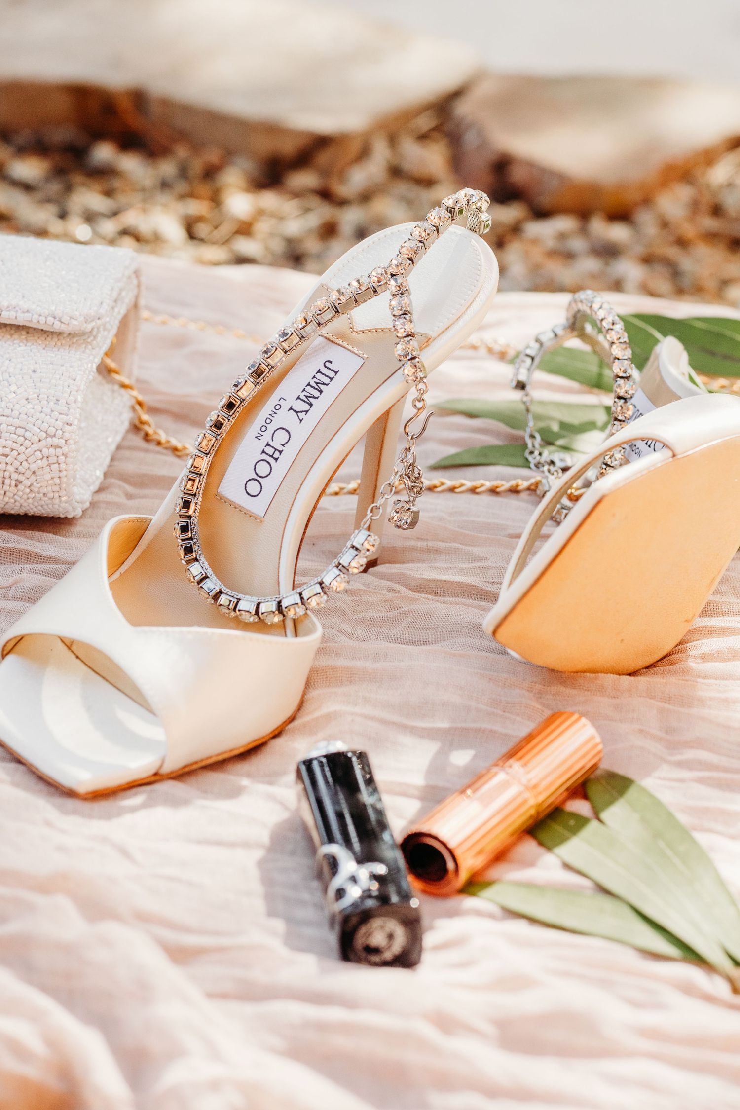 Elegant bridal shoes with rhinestone details and orange accessories arranged on white fabric with delicate green leaves.