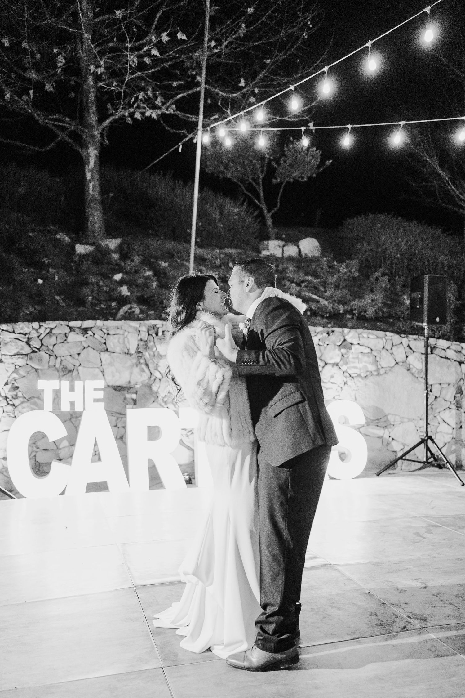 Black and white photo of a wedding couple dancing under string lights with illuminated letter decor.