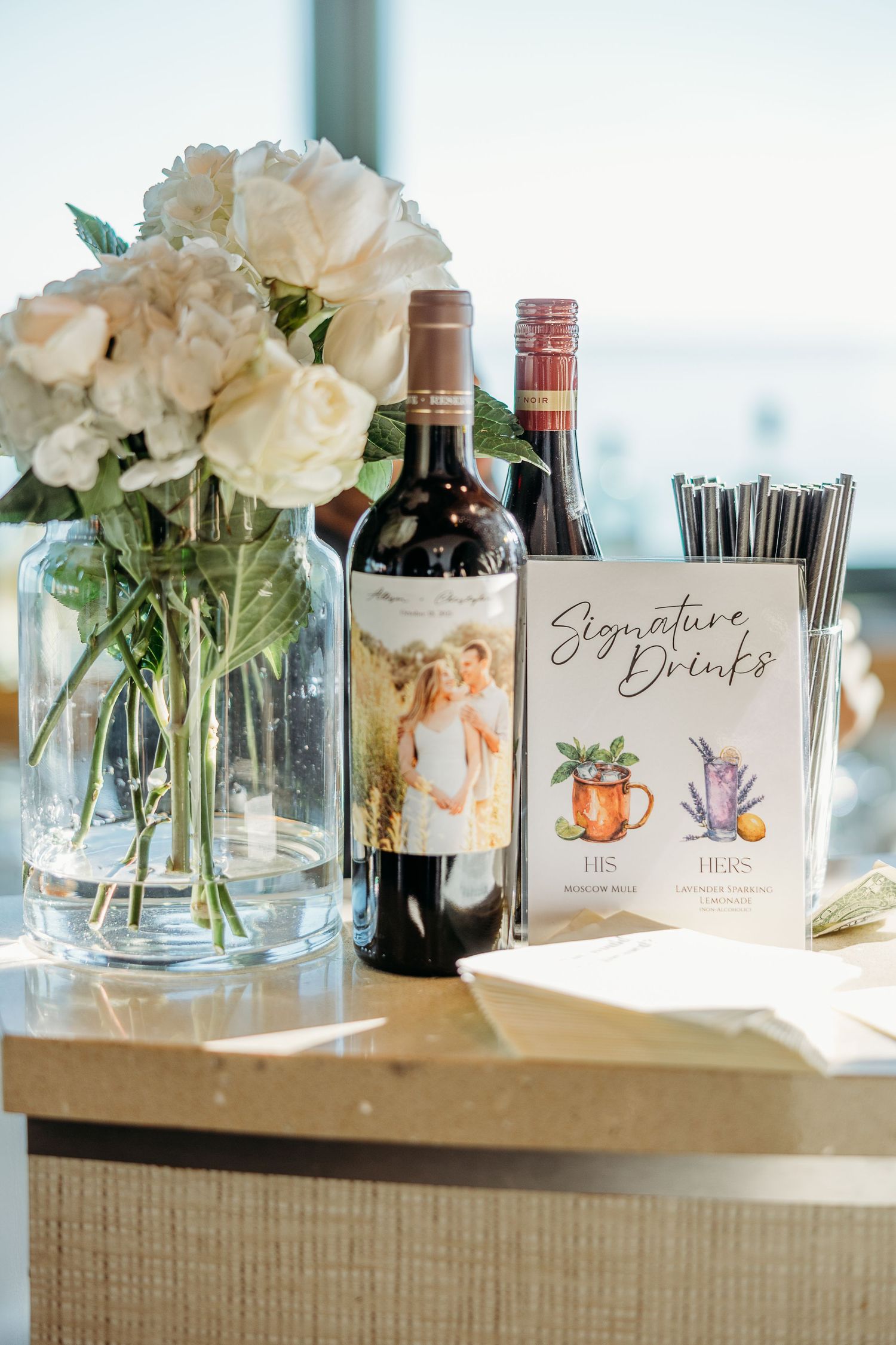 Wedding reception detail with white roses, wine bottles and signature drinks menu on wooden surface.