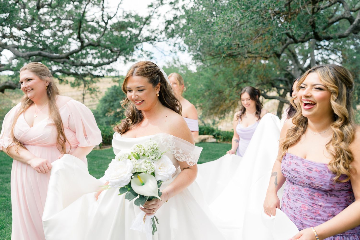 Smiling bride in off-shoulder white gown holds bouquet while walking with bridesmaids in pink dresses.