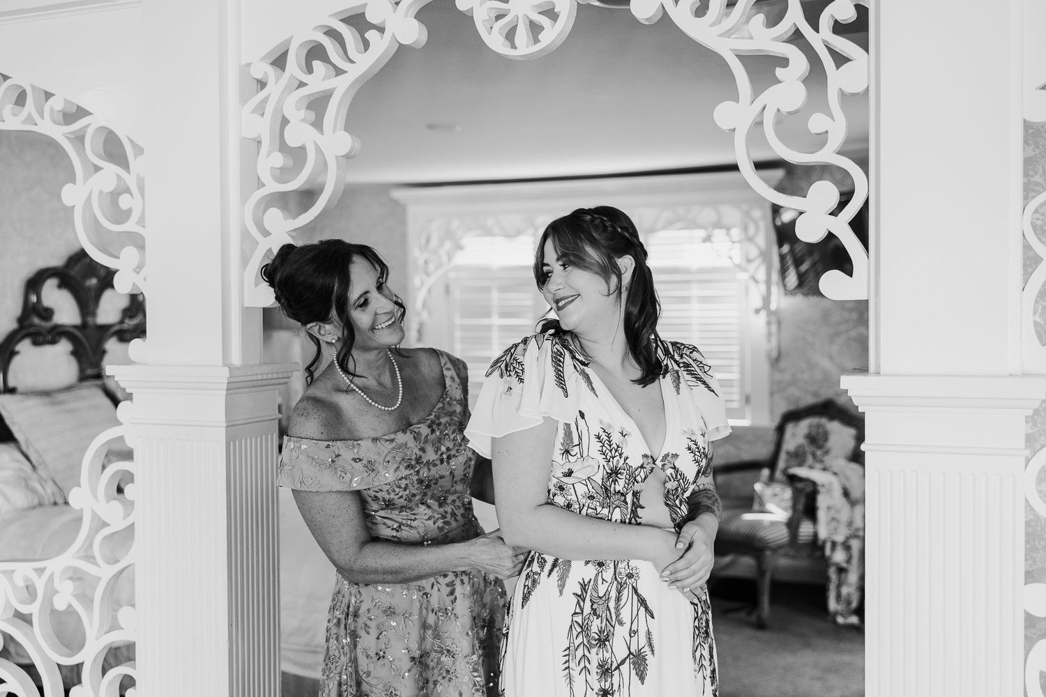Two women in floral dresses share a joyful moment under an ornate white arch during wedding preparations.
