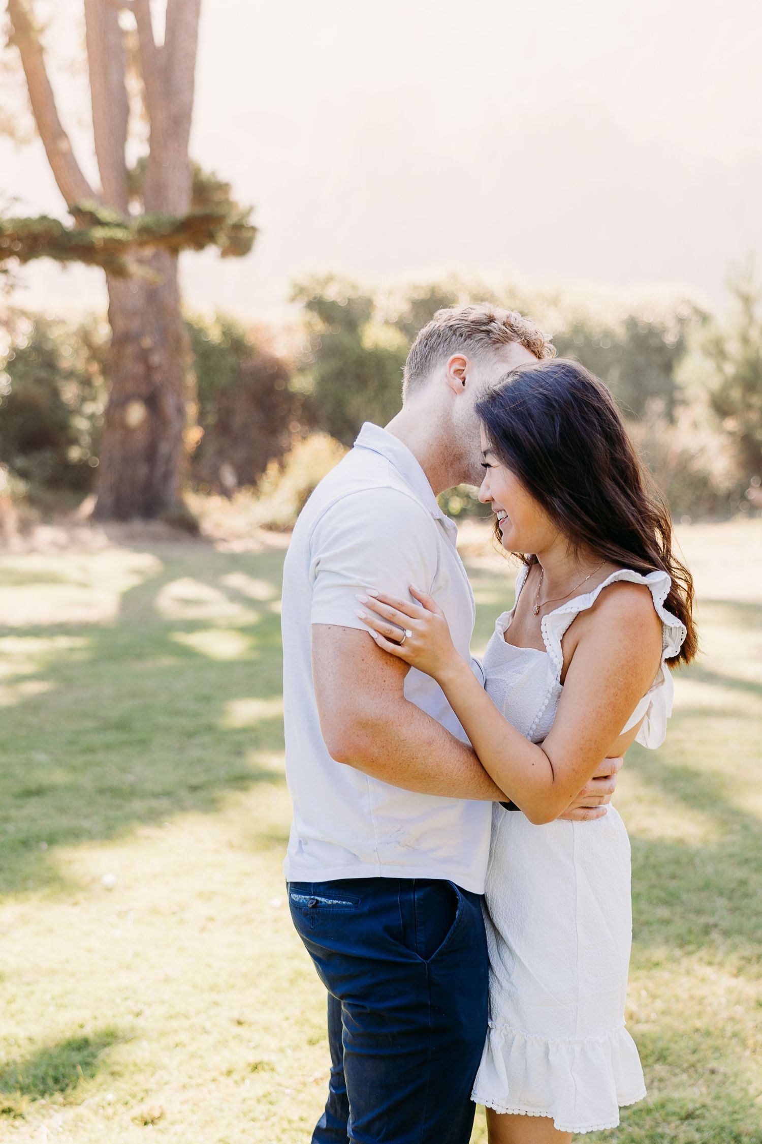 Couple laughing and embracing in a sunny park with trees in the background.