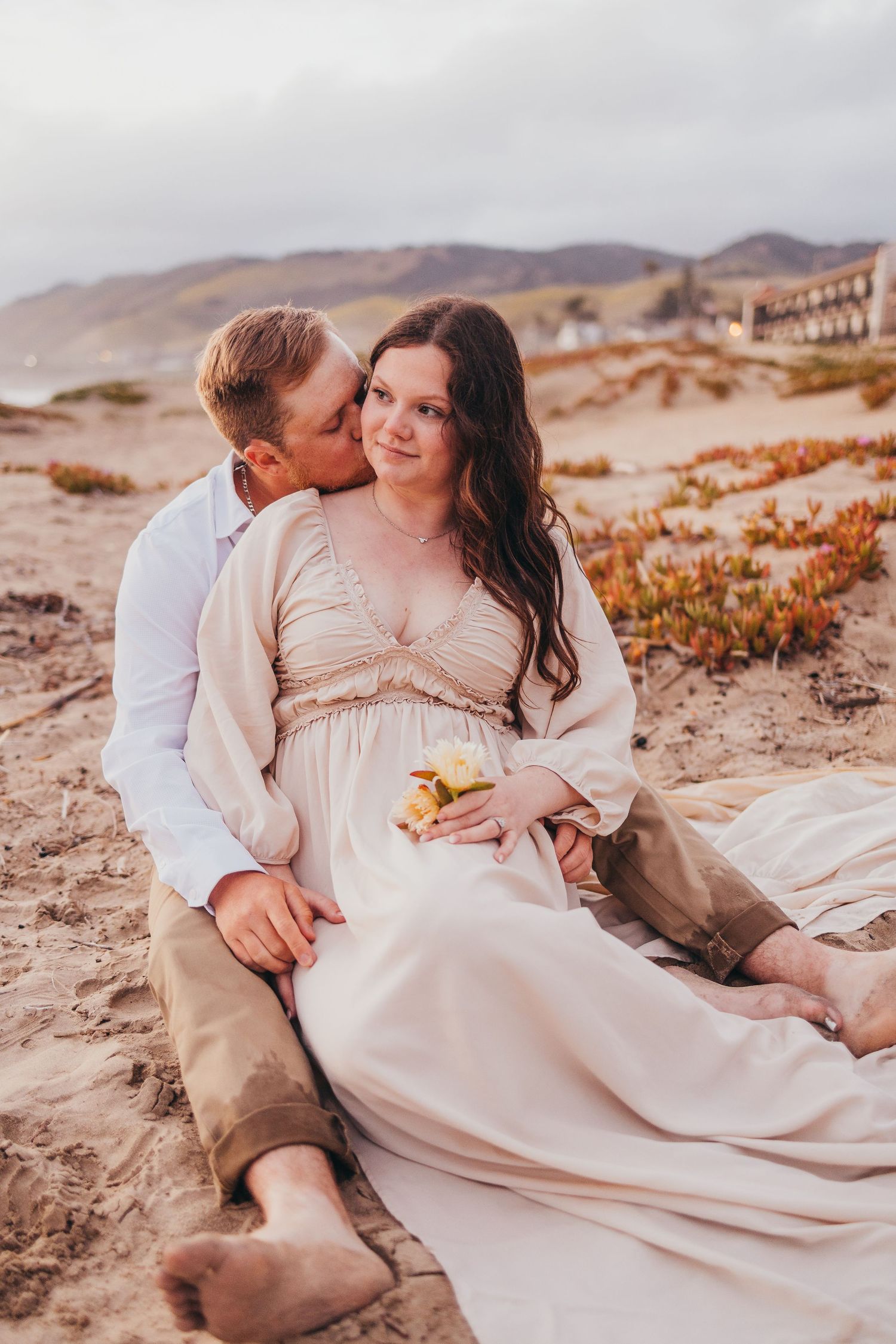 A couple in romantic embrace sits on sandy dunes during golden hour.