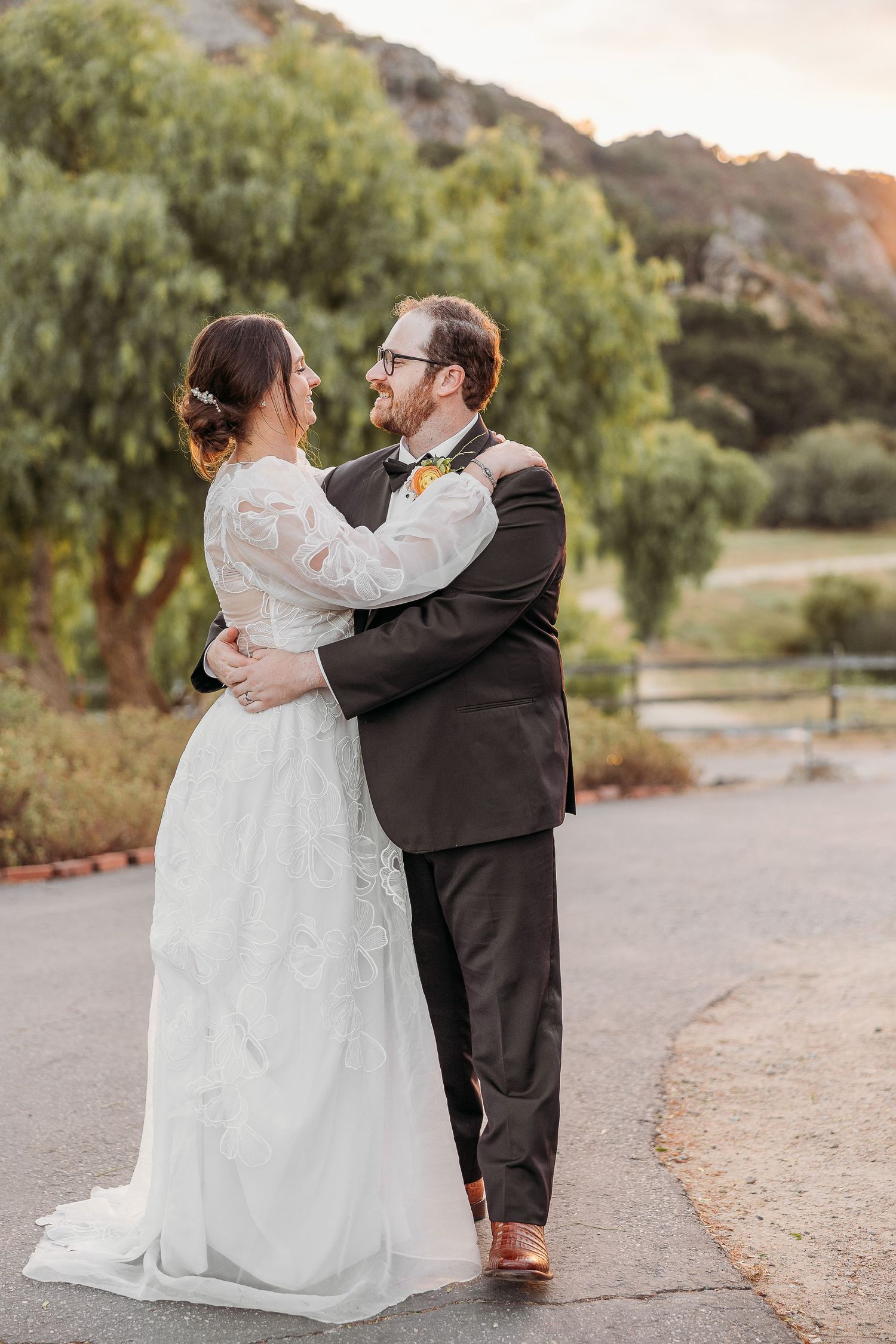 A couple shares a romantic moment in wedding attire on a path with willow trees and mountains in the background.