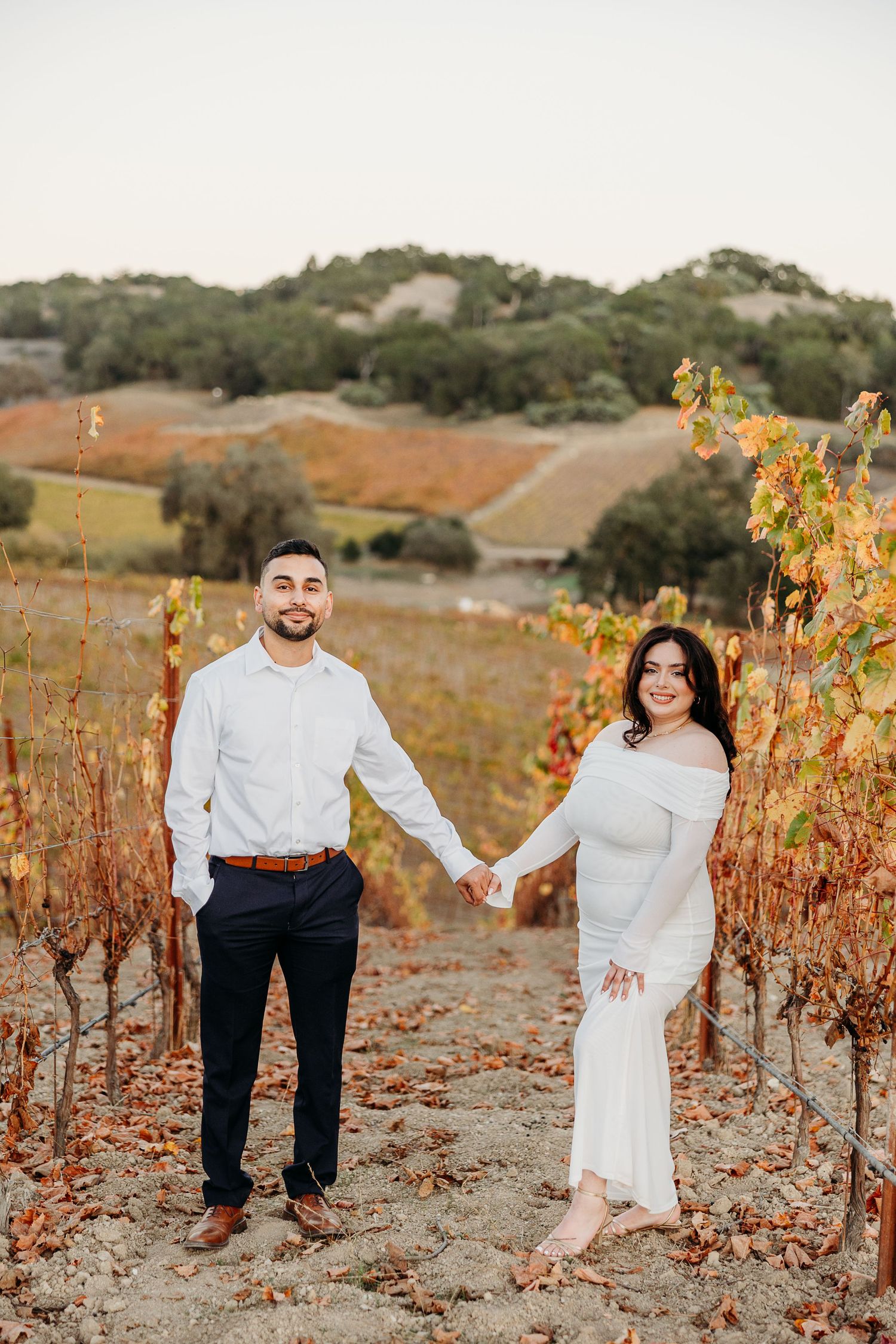 A couple poses playfully between rows of colorful fall grapevines in a rustic vineyard setting.