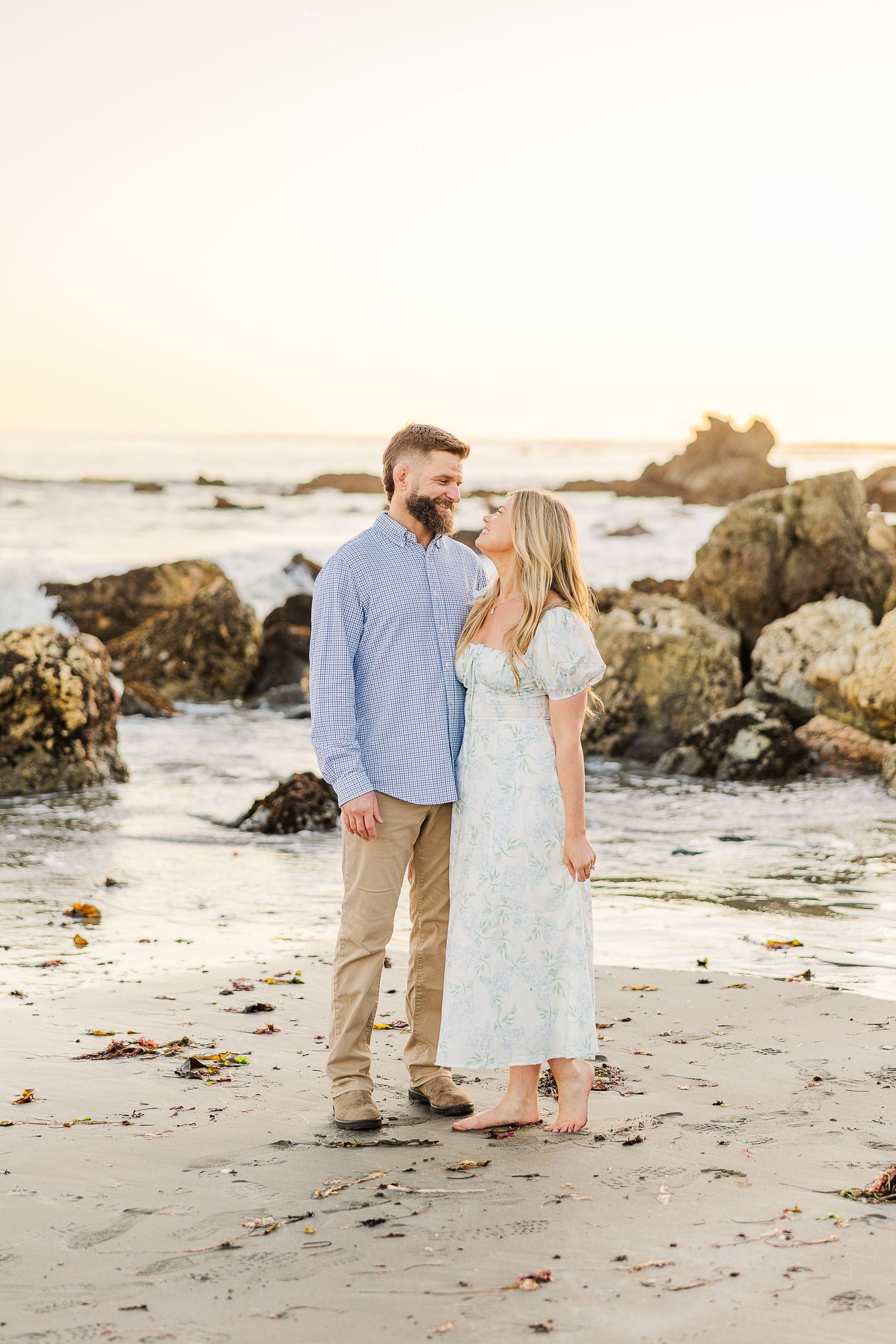 Couple shares an intimate moment on a rocky beach at sunset wearing coordinated blue and white outfits.
