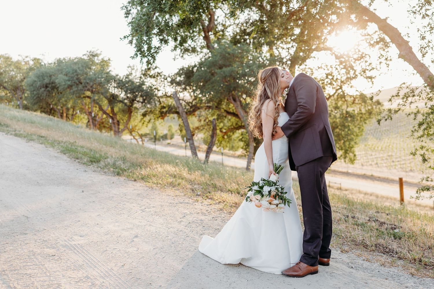 Wedding couple walks hand in hand along a scenic pathway with natural stone walls and trees during golden hour.