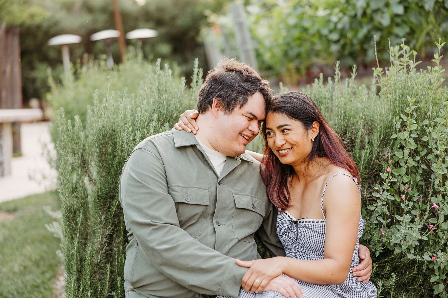 A couple embraces while sitting outdoors surrounded by green plants and natural foliage in bright sunlight.