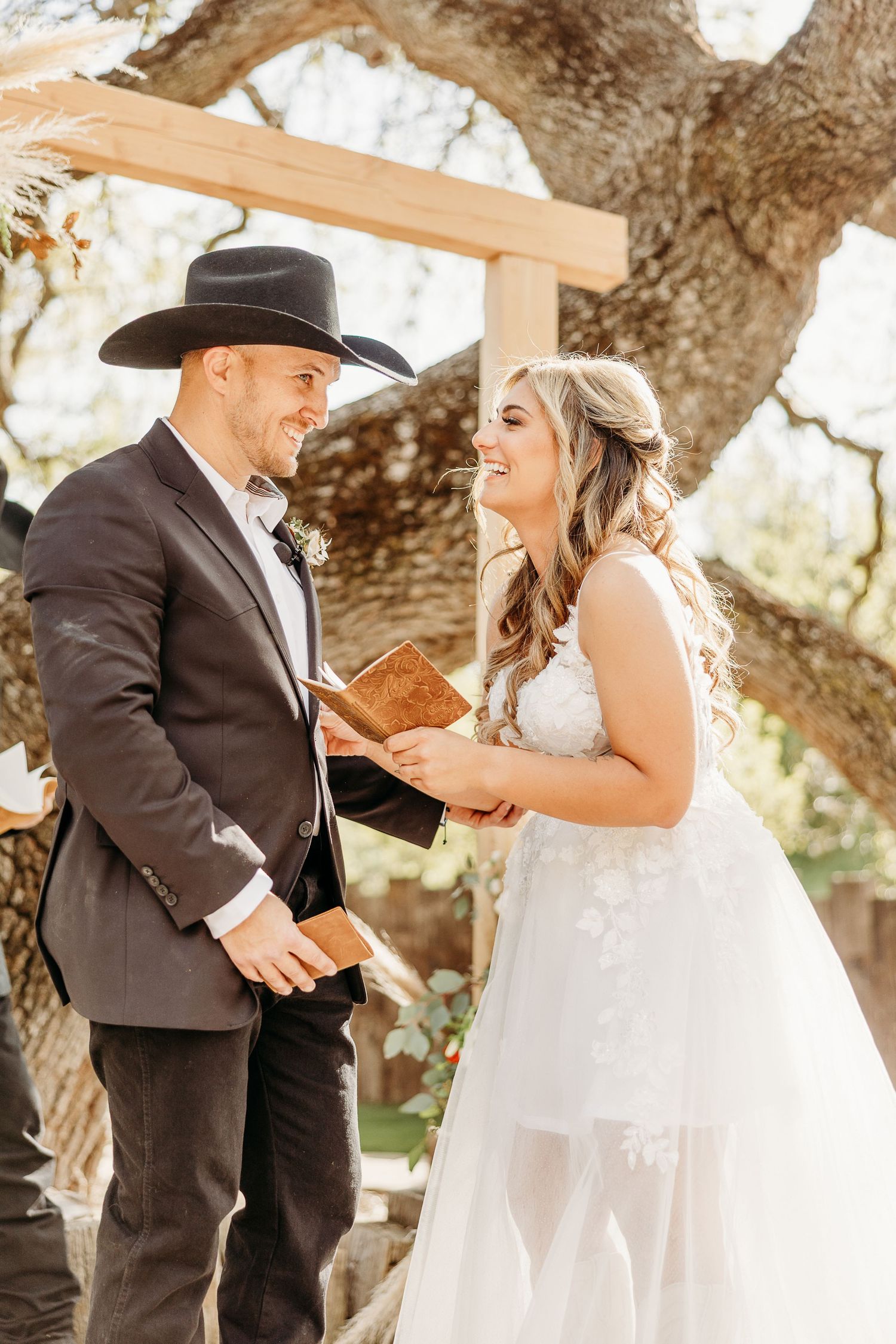 A bride in a white gown and groom in western wear share a joyful moment under an oak tree arch during their ceremony.