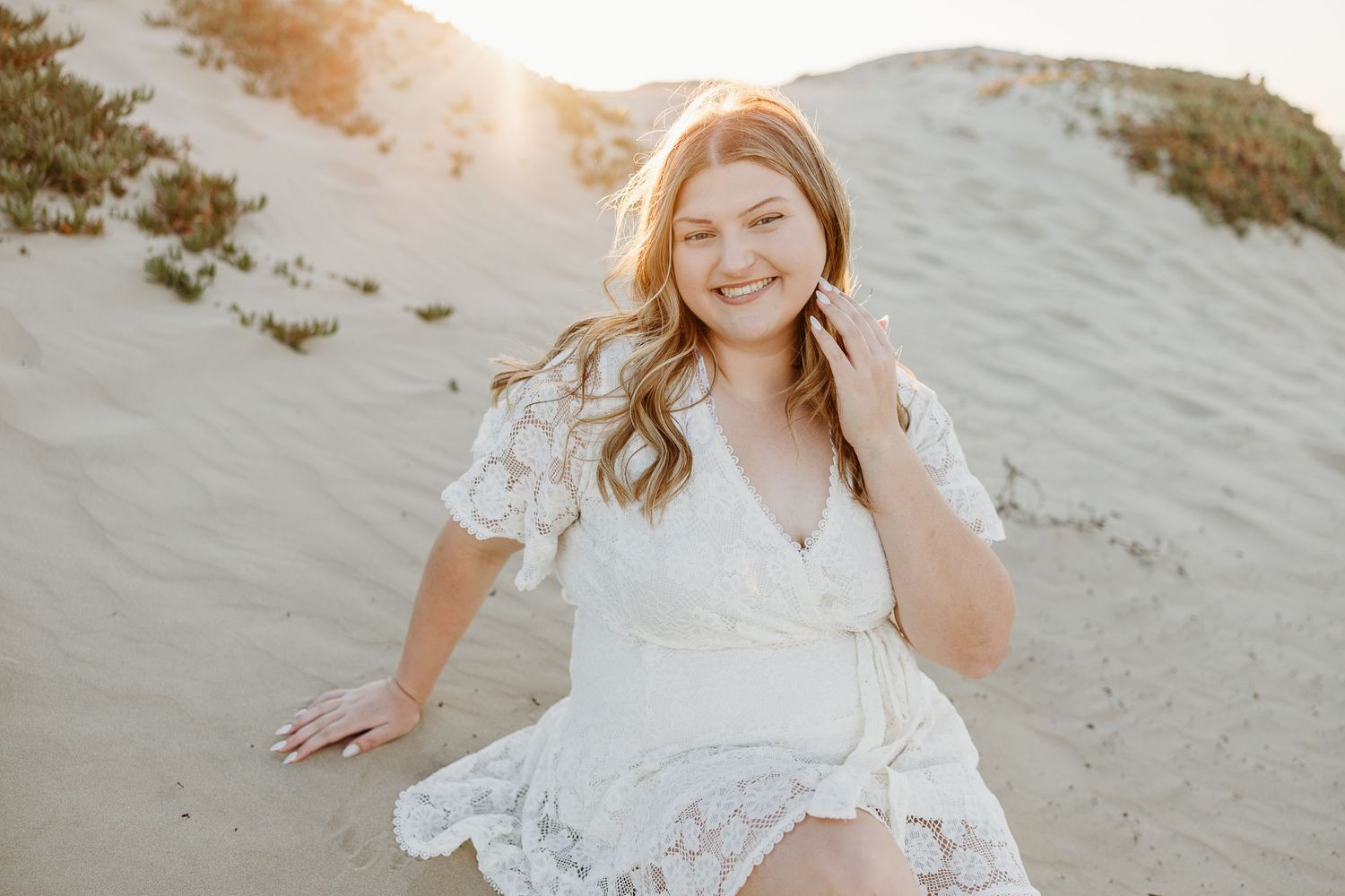 Graduate in white lace dress sits gracefully on sandy dunes during golden hour photoshoot.