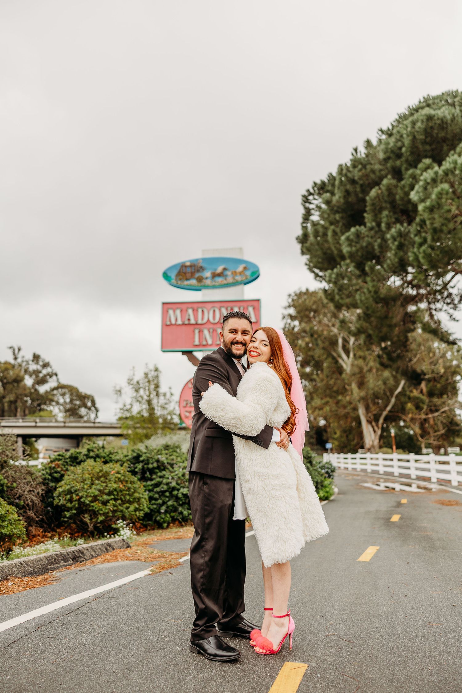 A couple embraces on a roadway near vintage signage and lush greenery in a romantic outdoor setting.