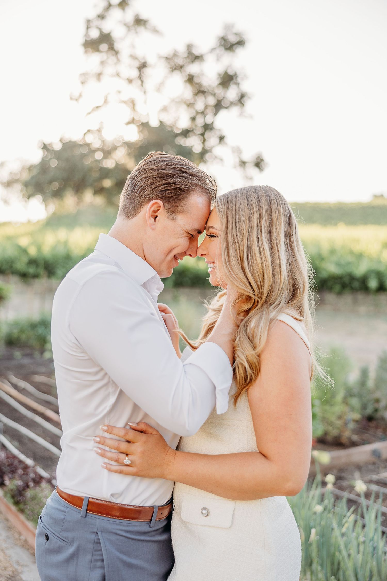 A romantic couple embraces in a garden setting during golden hour, dressed in light summer attire.