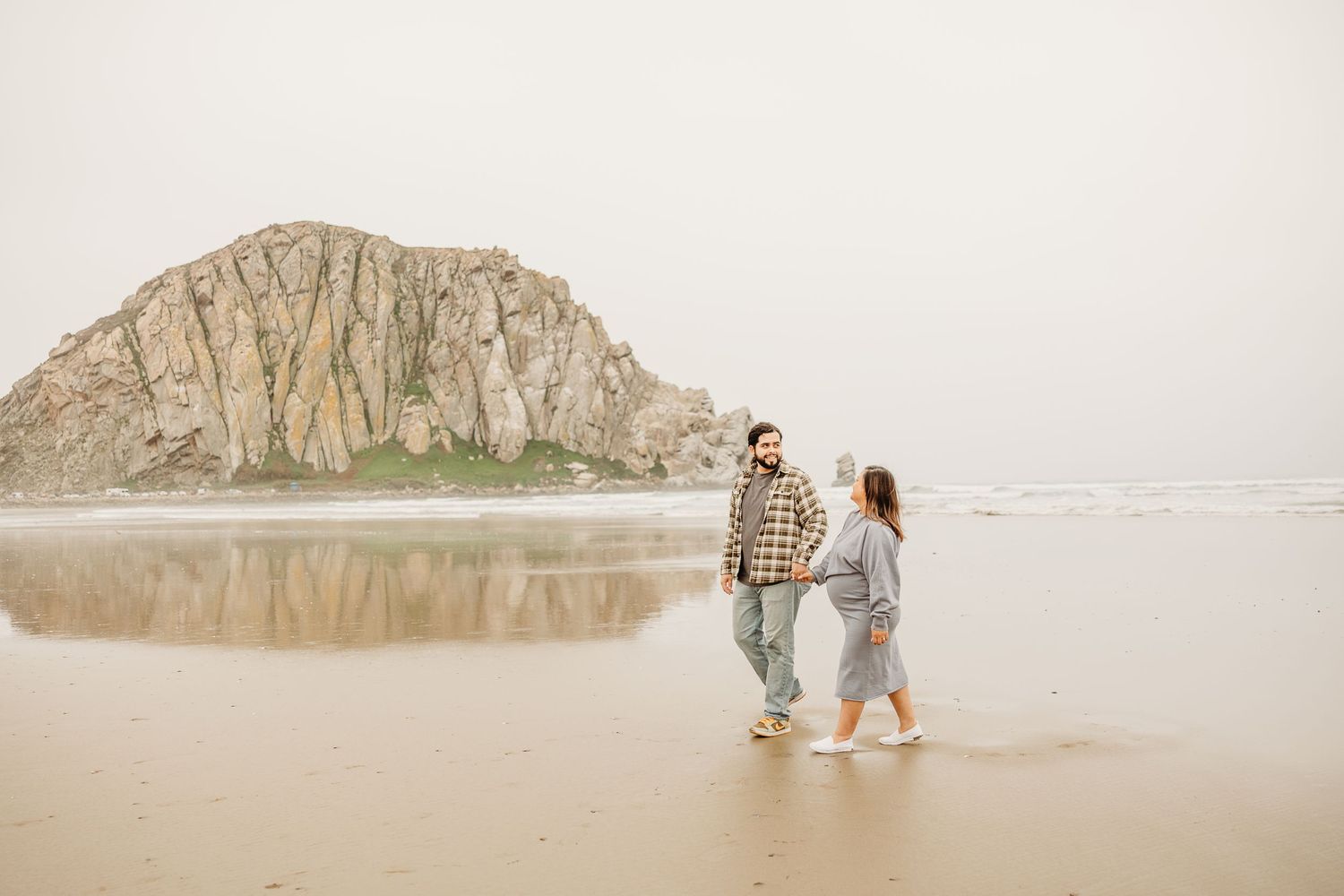 Couple walking along foggy beach with Morro Rock formation visible in background.