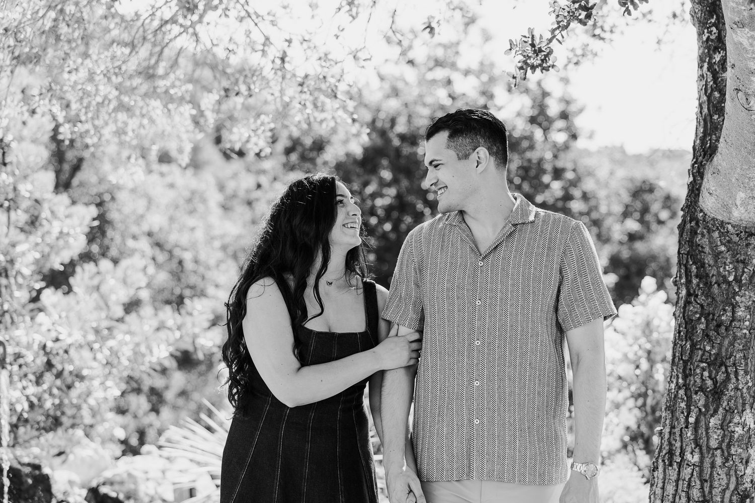 A romantic couple poses together under shady trees in a natural outdoor setting.