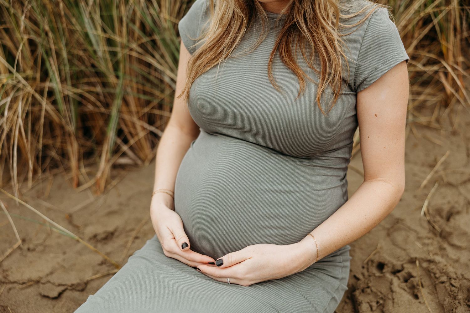 A pregnant woman in a grey dress sits among tall grass while gently cradling her baby bump.
