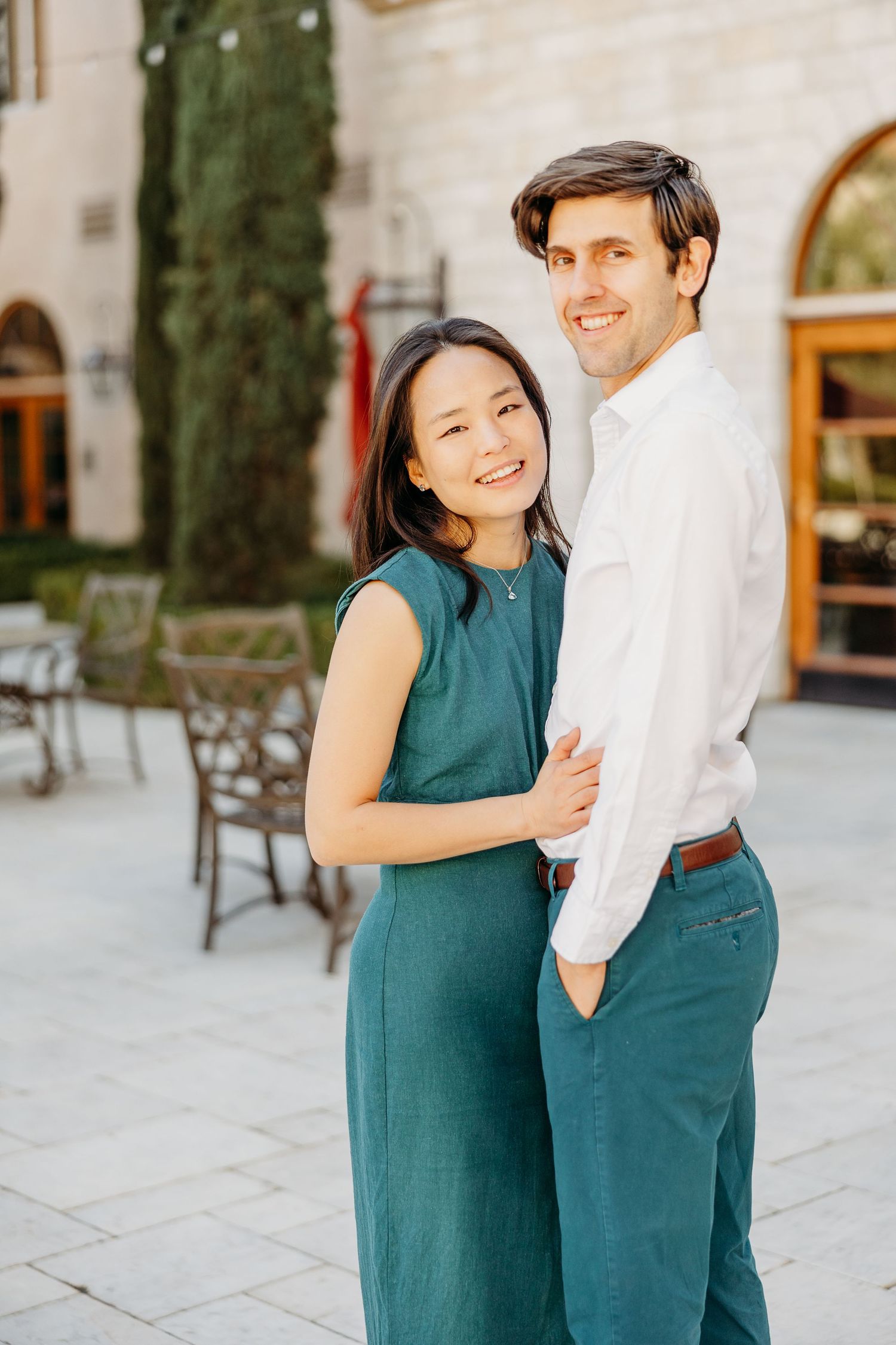 A couple in coordinating teal and white outfits embrace and laugh together on a sunny patio.