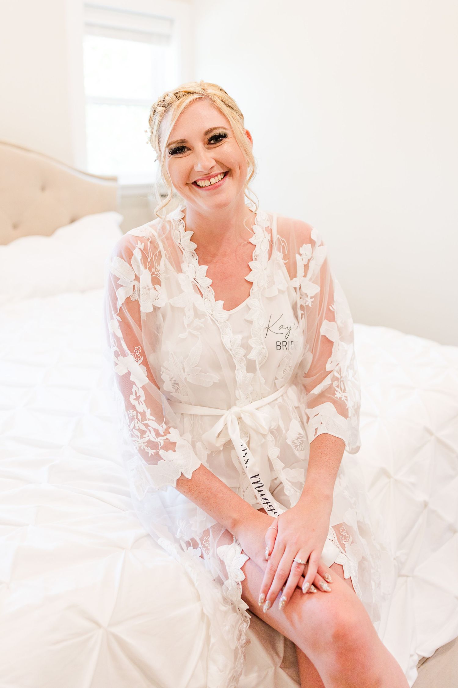 Person wearing white lace robe smiles while sitting on bed during wedding preparations.