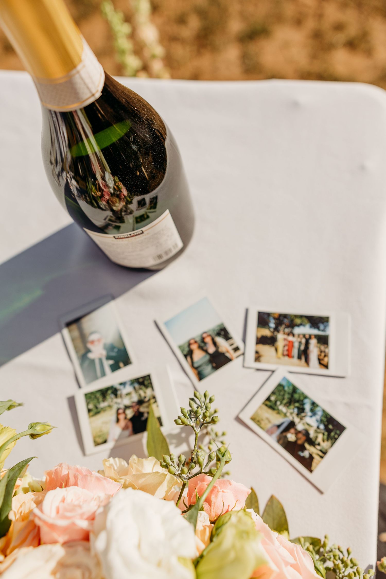 Wedding reception table with champagne bottles, pink roses, and scattered polaroid photos on white tablecloth.