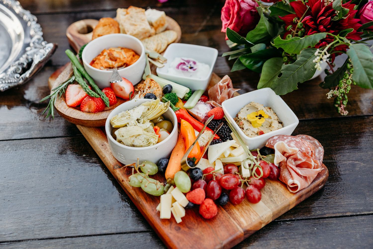 A wooden charcuterie board filled with meats, cheeses, fruits and dips alongside red roses.