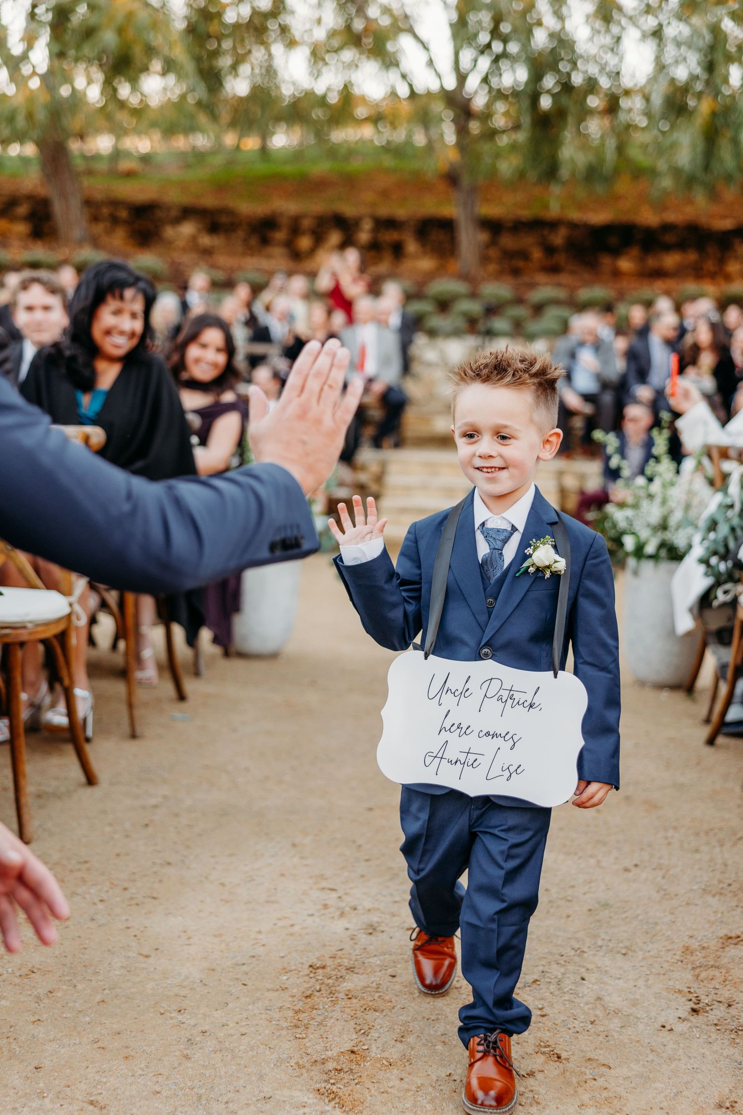 A young ring bearer in a navy suit gives a high five while walking down the aisle.