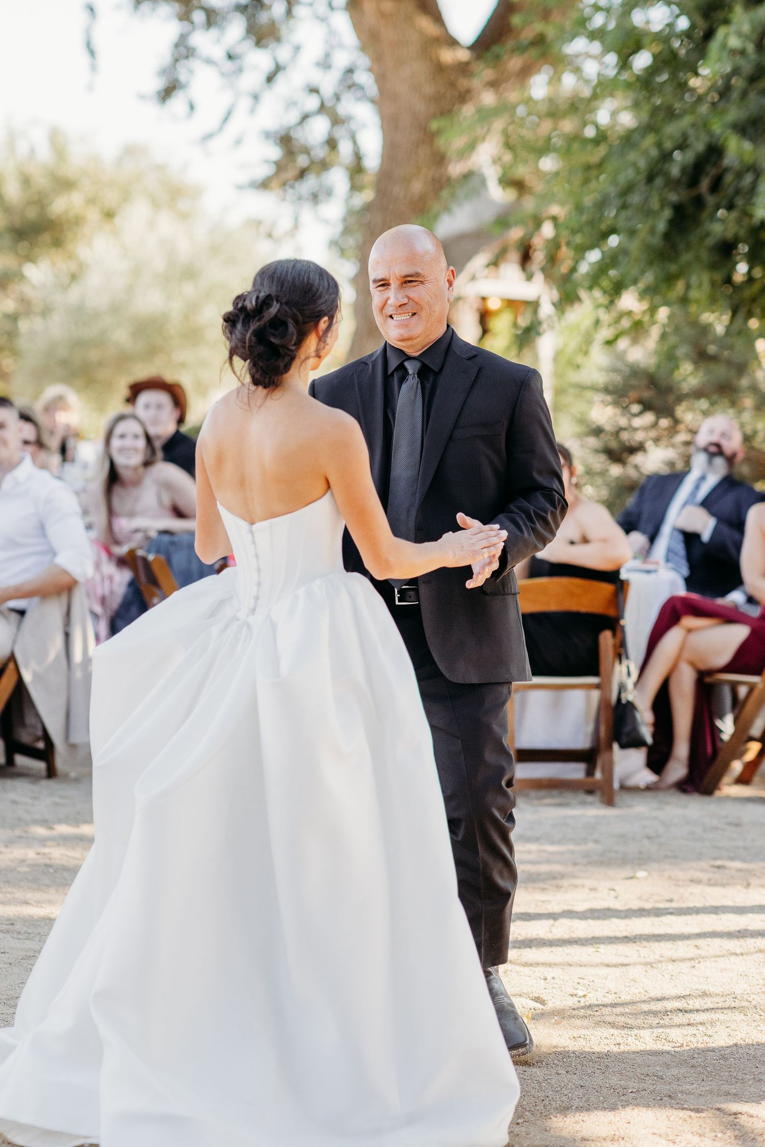 Wedding ceremony guests seated outdoors under trees watching couple share their first dance.
