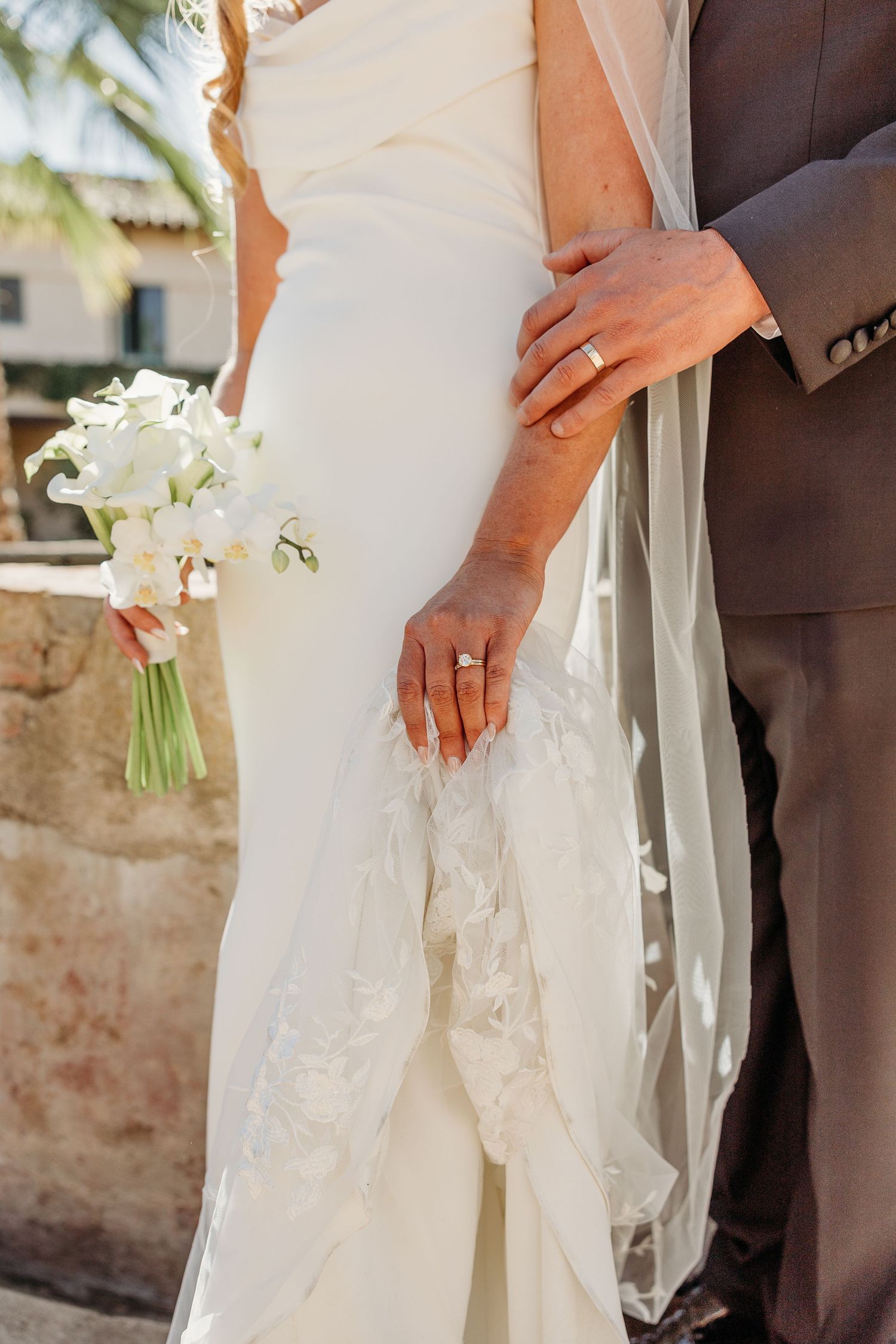 Close-up of wedding dress details with white orchid bouquet and hands showing wedding rings.