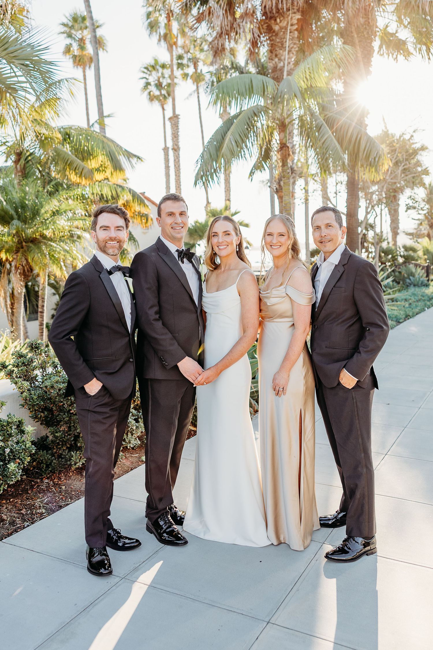 Wedding party in formal attire posing outdoors with palm trees and sunlight in the background.