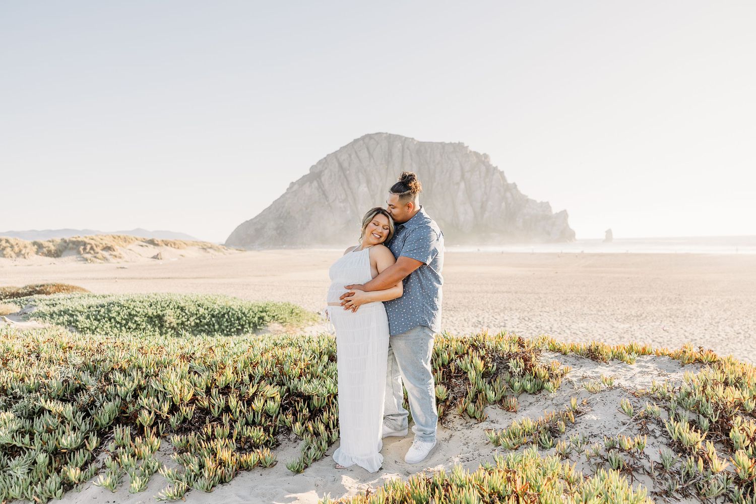 A romantic couple embraces on a scenic beach with mountains and wildflowers at sunset.