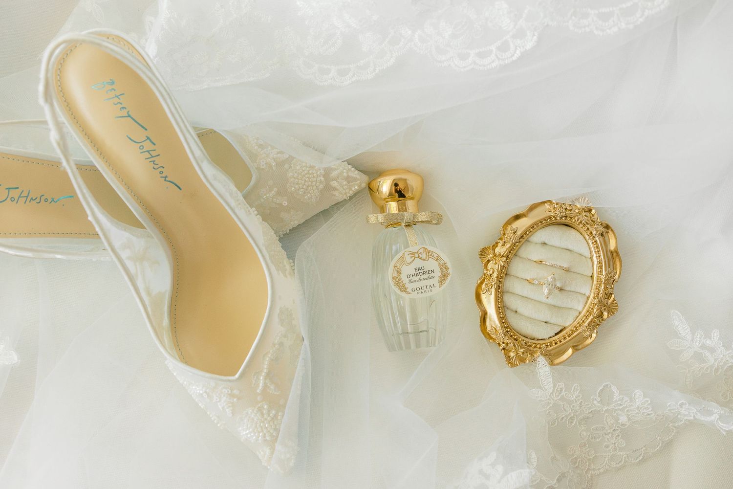 White lace wedding heels, perfume bottle, and gold ring dish arranged on bridal veil fabric.