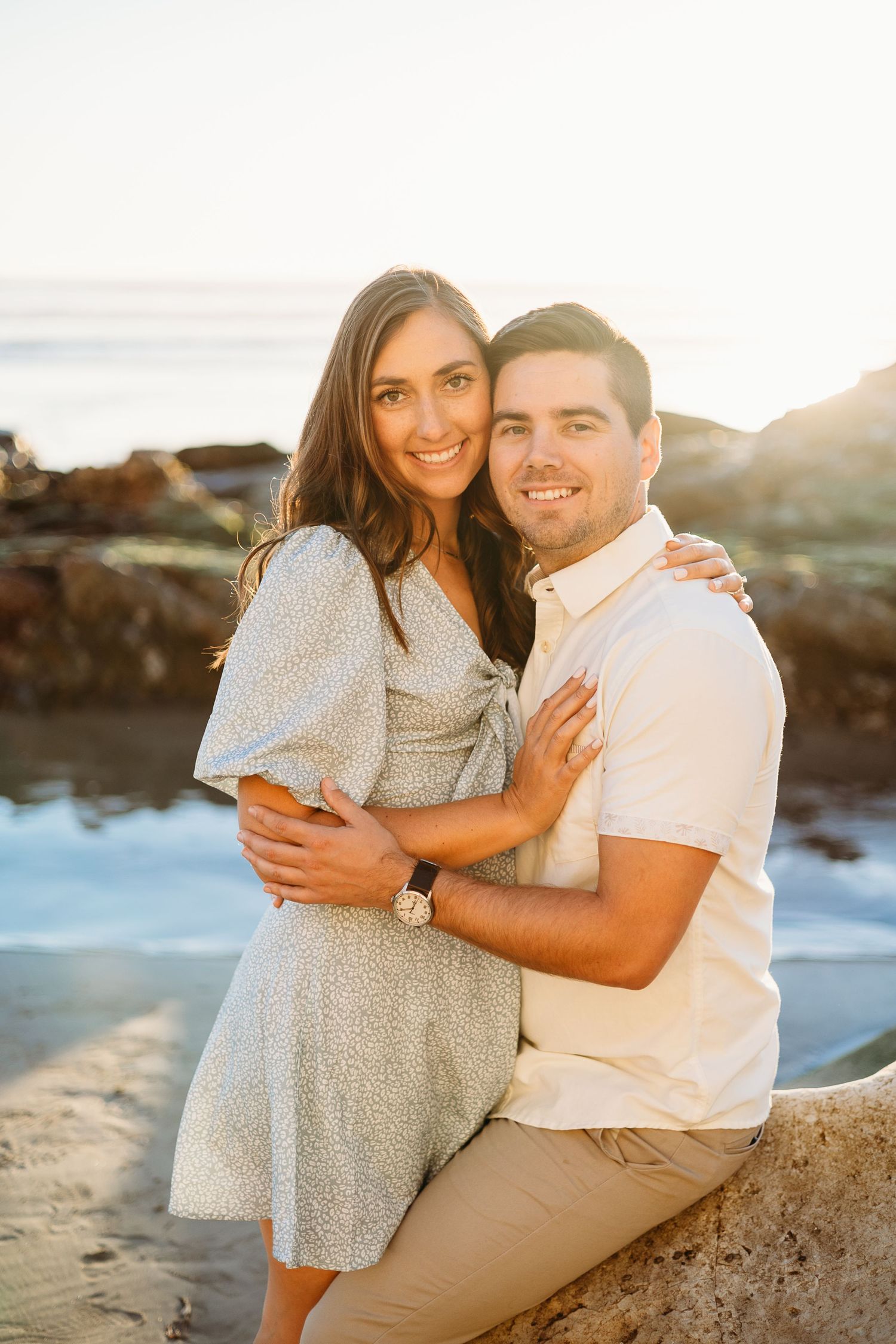 A couple embraces on a rocky beach during golden hour with ocean waves in the background.