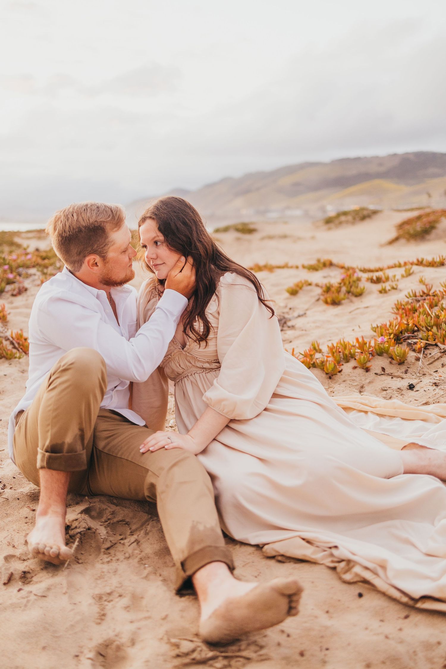Romantic coastal photoshoot with couple nestled together on sandy dunes.