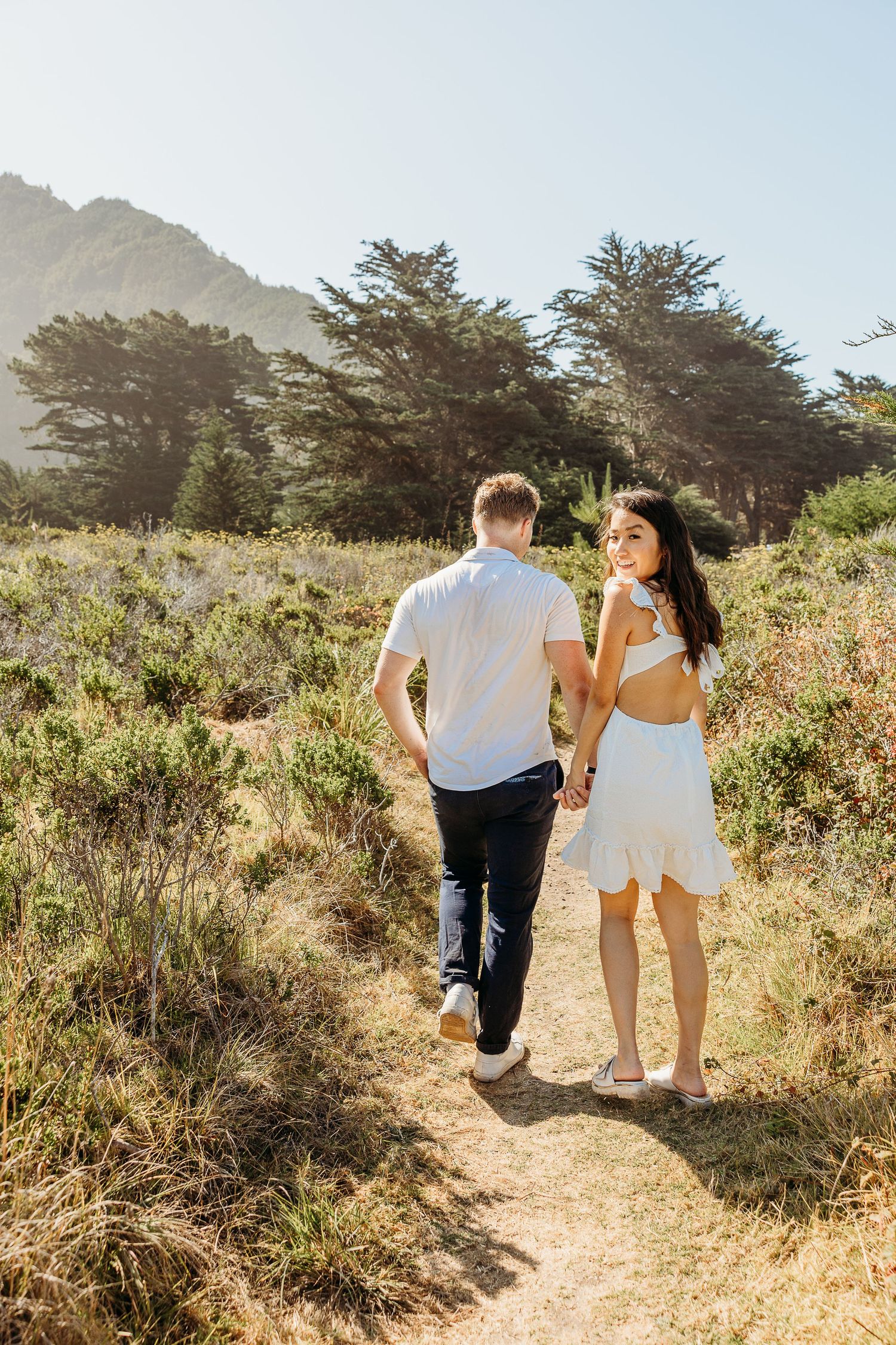 Couple in white and dark outfits walking along a sunny coastal trail.