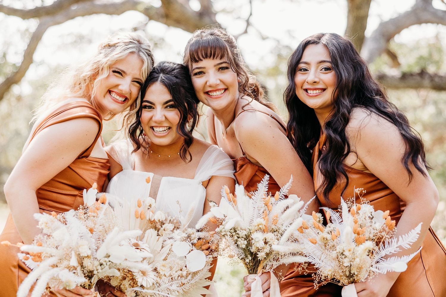 Bridal party in rust-colored dresses share a candid laugh while holding cream-colored bouquets.