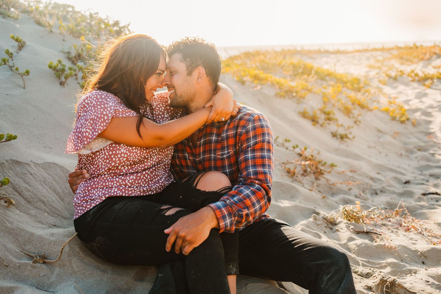 Romantic couple sharing an intimate moment on sandy dunes at sunset in matching plaid and floral outfits.