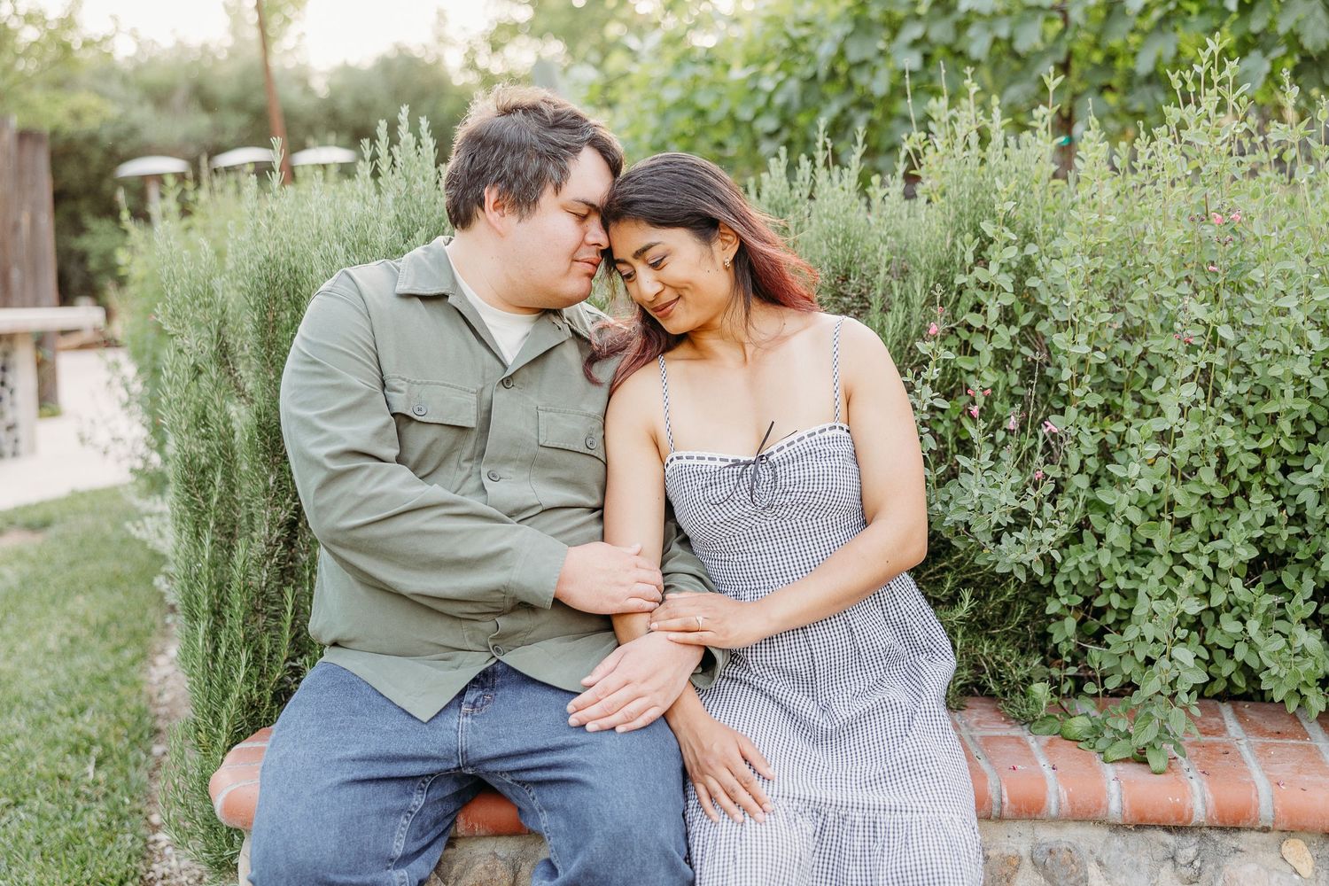 A couple shares an intimate moment while sitting together on a brick wall surrounded by lush garden greenery.