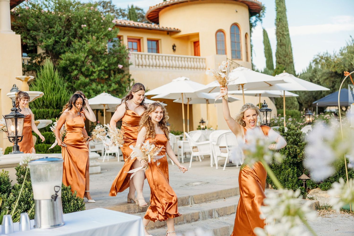 Wedding party in flowing copper-colored dresses descending stone steps at an elegant outdoor venue.