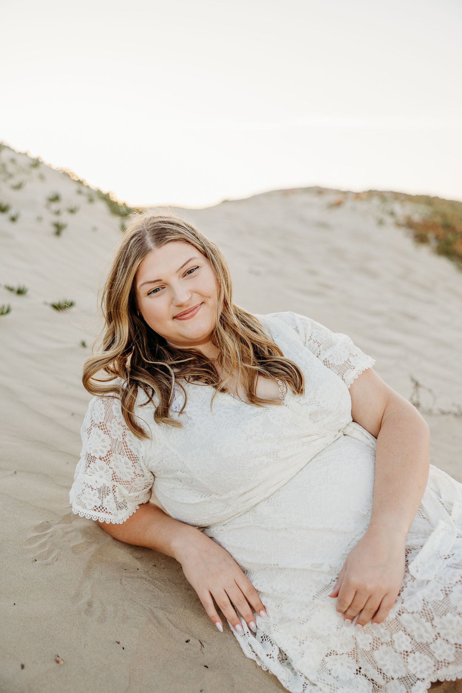 Someone in a white lace dress poses on sandy dunes during a beach photoshoot.