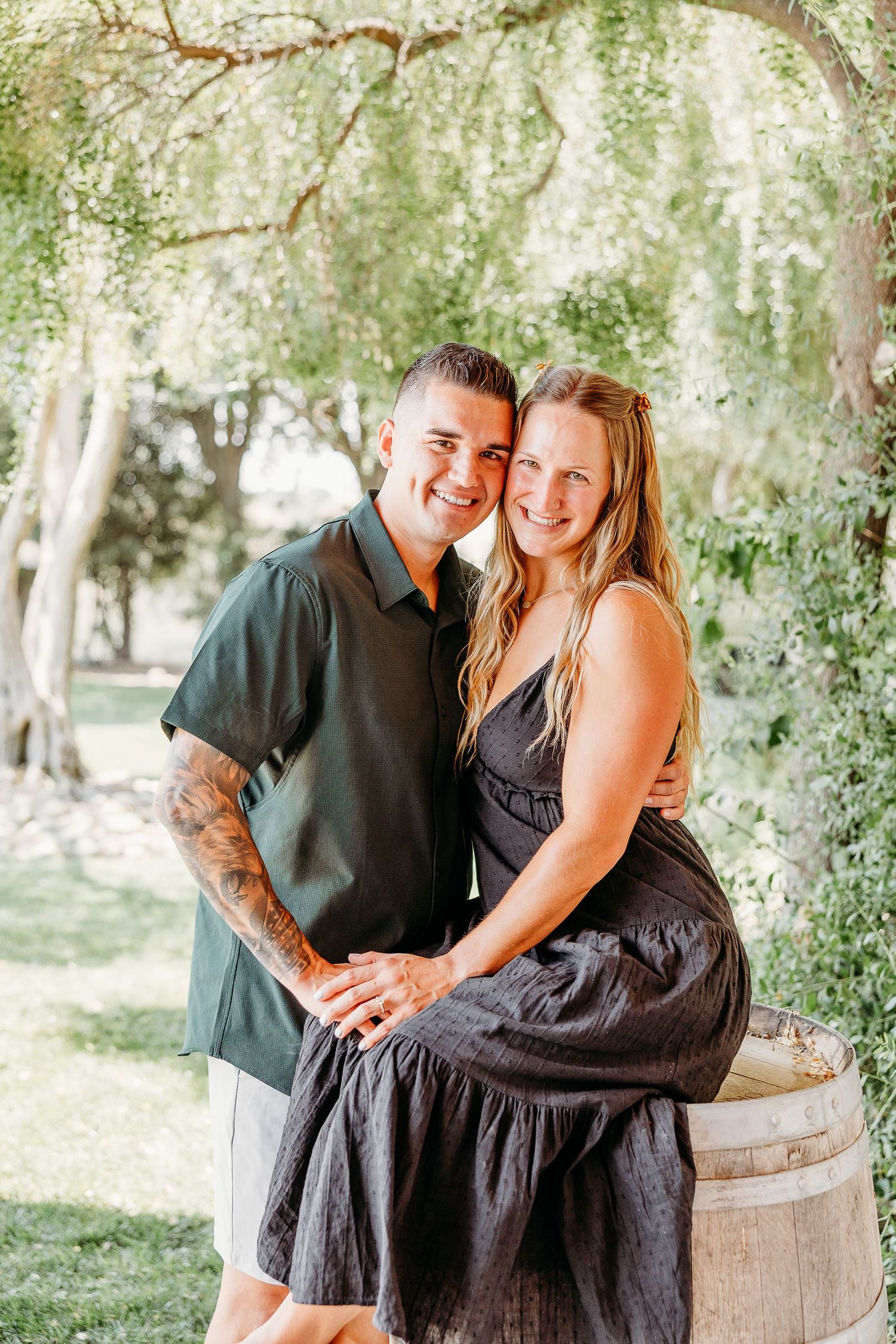 A couple poses playfully together near wooden barrels in a garden setting with trees in the background.